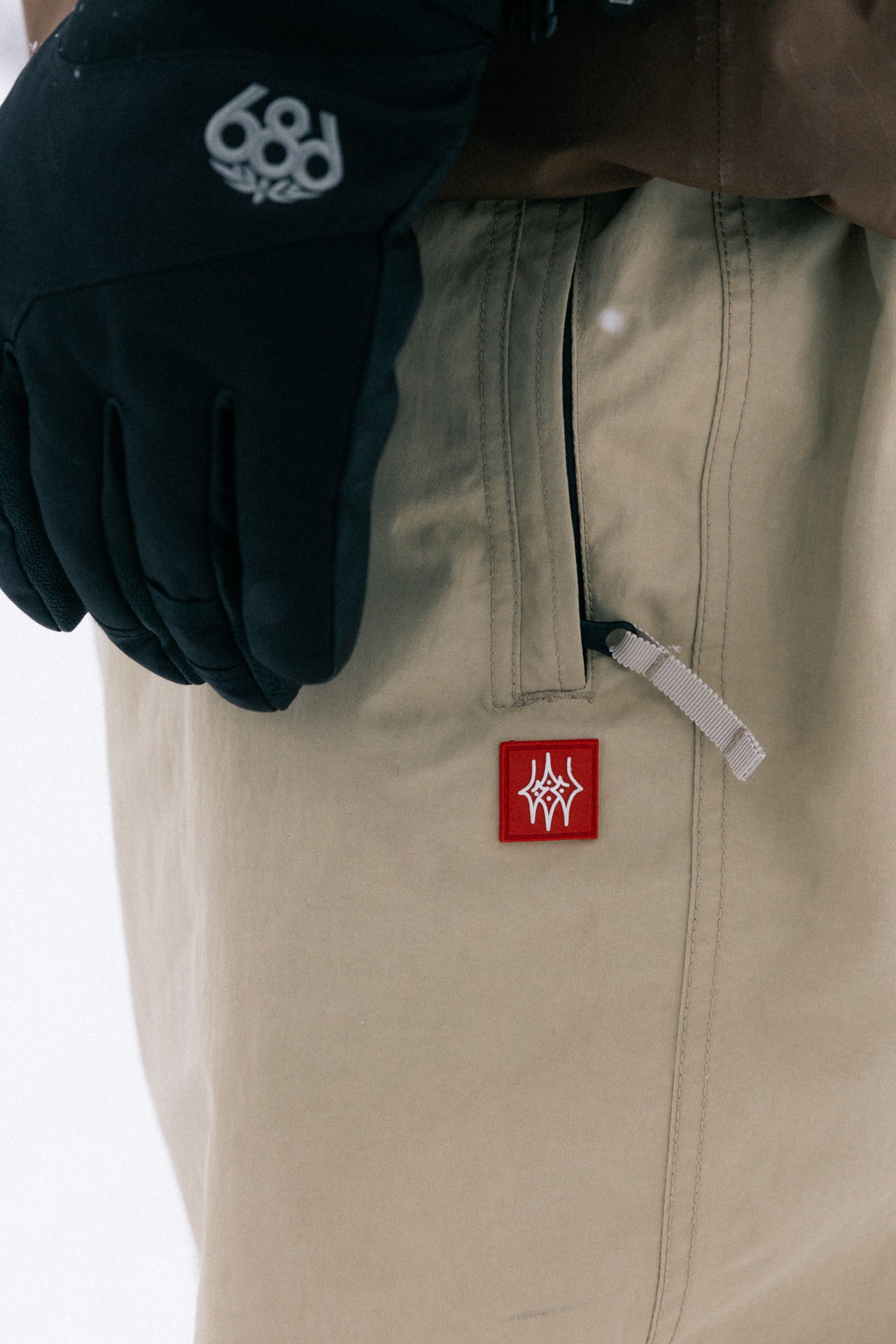 A person wearing the 686 Men’s 686 Dojo® Shell Pant in beige, featuring a visible red patch and white drawstring, stands with their hand near the pocket. They are also wearing black gloves and a brown jacket sleeve from brand 686.