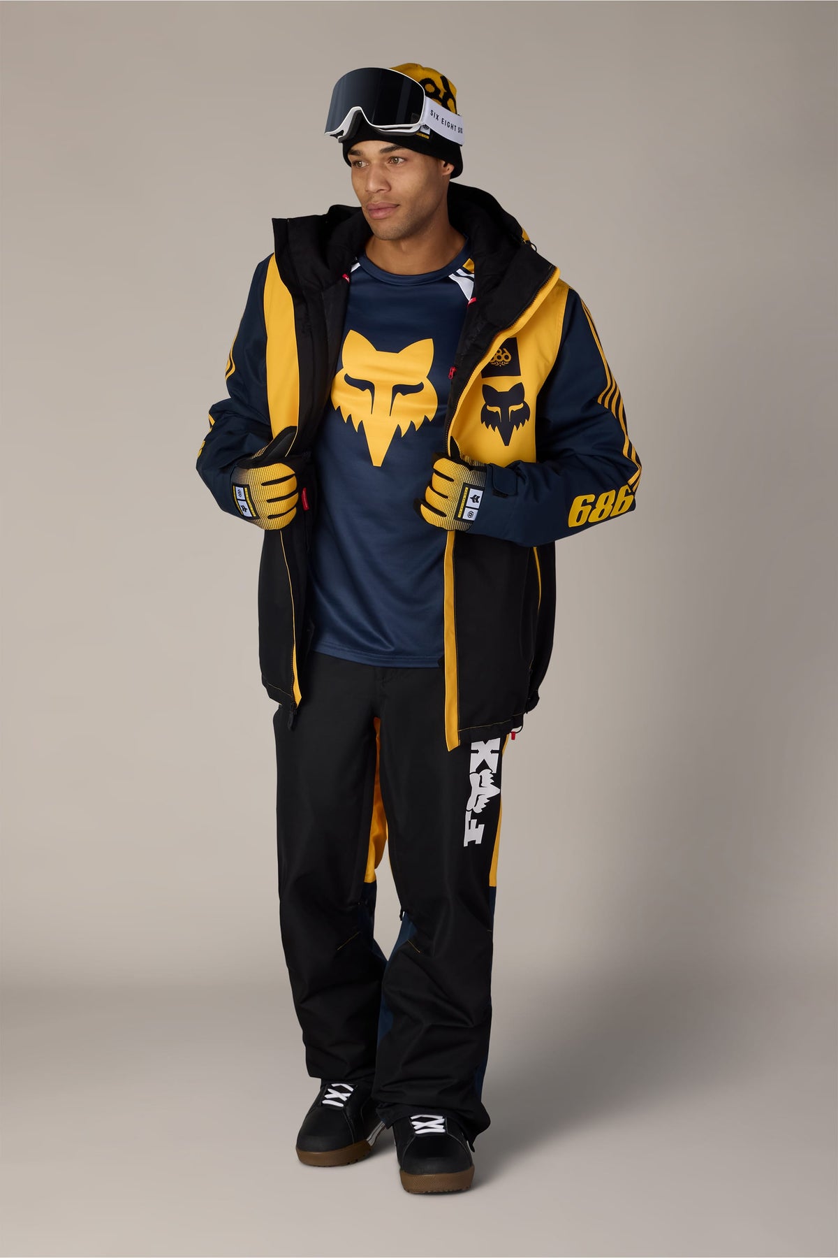 A man wearing a Fox Racing beanie and goggles, a navy and yellow snowboarding jacket, and 686 Men’s Fox Racing Spec Shell Pant.