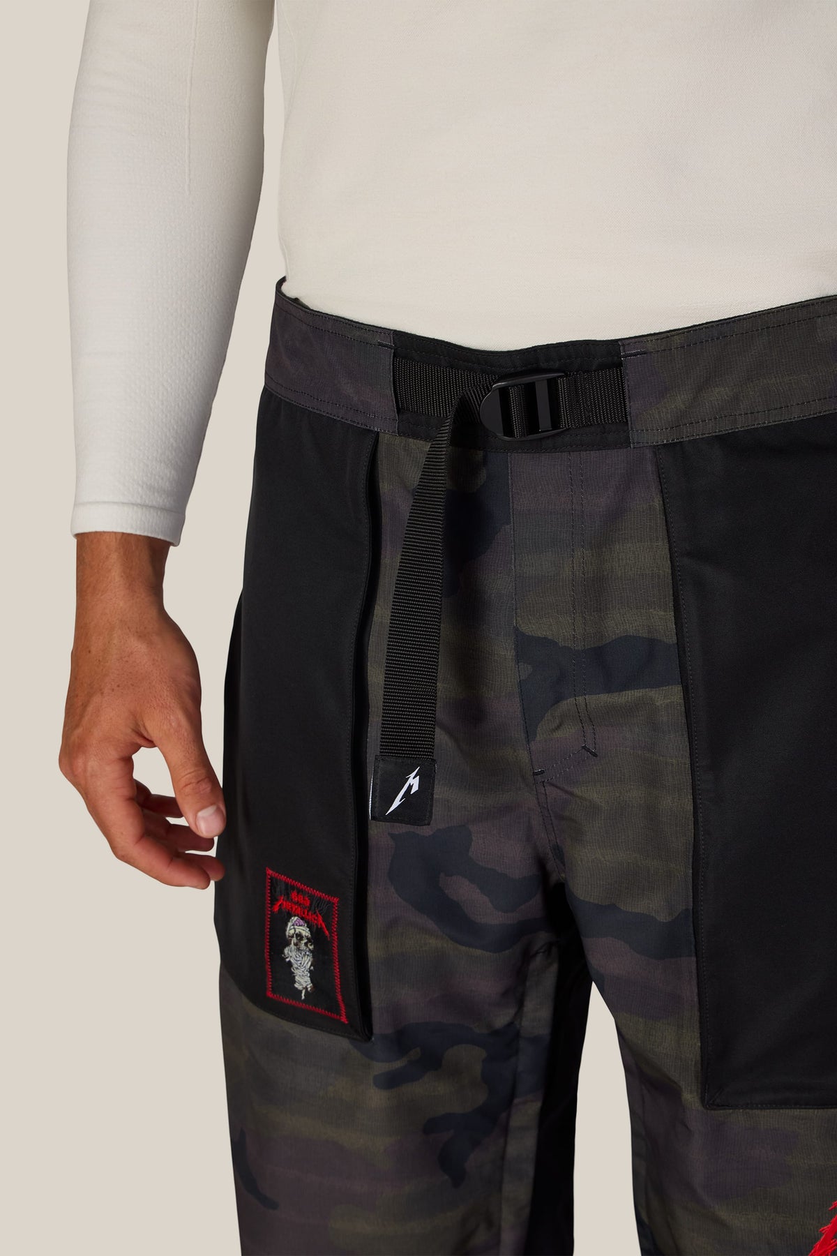 Close-up of 686 Men’s Metallica One Shell Pant in camouflage, featuring a black integrated webbing belt with a buckle and a square patch with red trim and white graphic detailing on the right thigh.