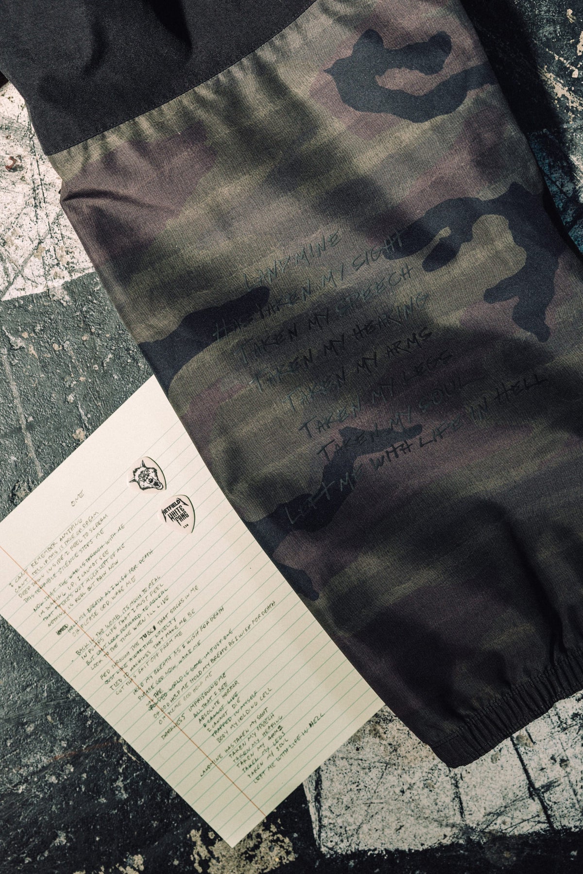 686 Men’s Metallica One Shell Pant in camo-print waterproof polyester featuring lyrics from the song One, displayed next to a handwritten lyric sheet on lined paper.