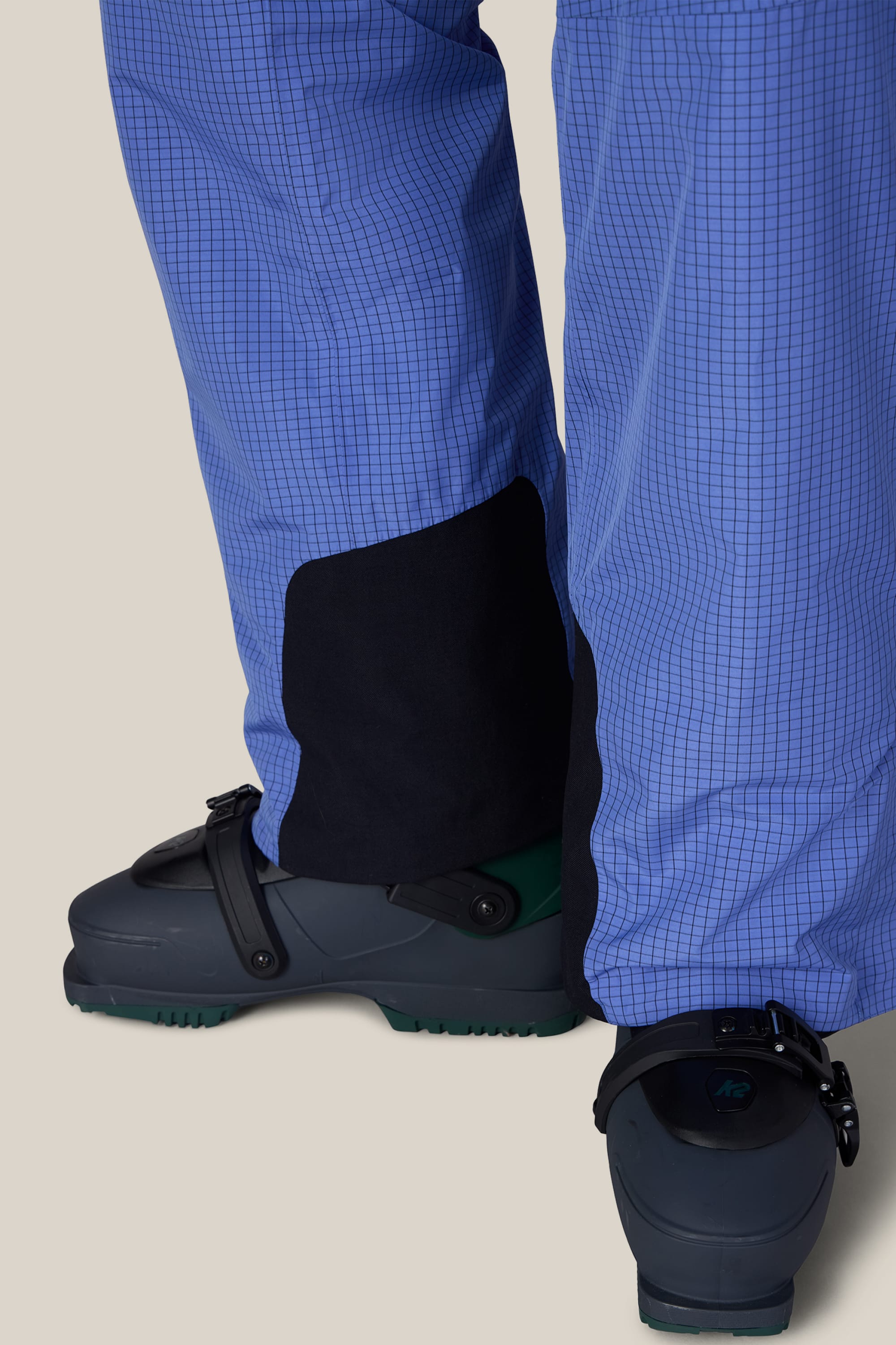 Close-up of a person wearing 686 Men’s Hydra™ Thermagraph® Pant in a blue grid pattern with black gaiters over dark ski boots, set against a light background.