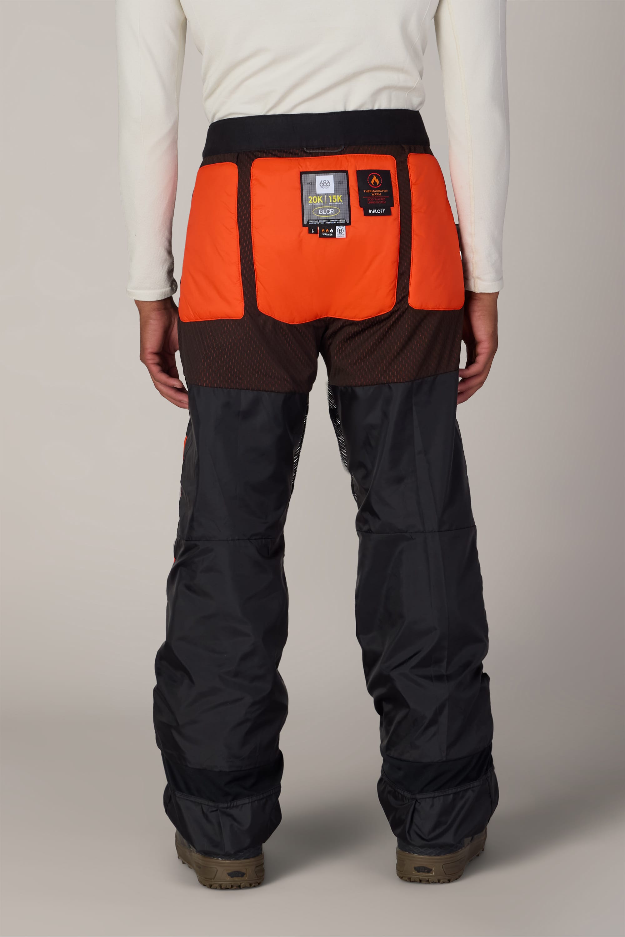 A person is standing with their back to the camera, wearing 686 Men’s Hydra™ Thermagraph® Pant by 686, featuring black polyester fabric with bright orange panels on the upper back and mesh details. They are also wearing brown outdoor boots and a white long-sleeve shirt.