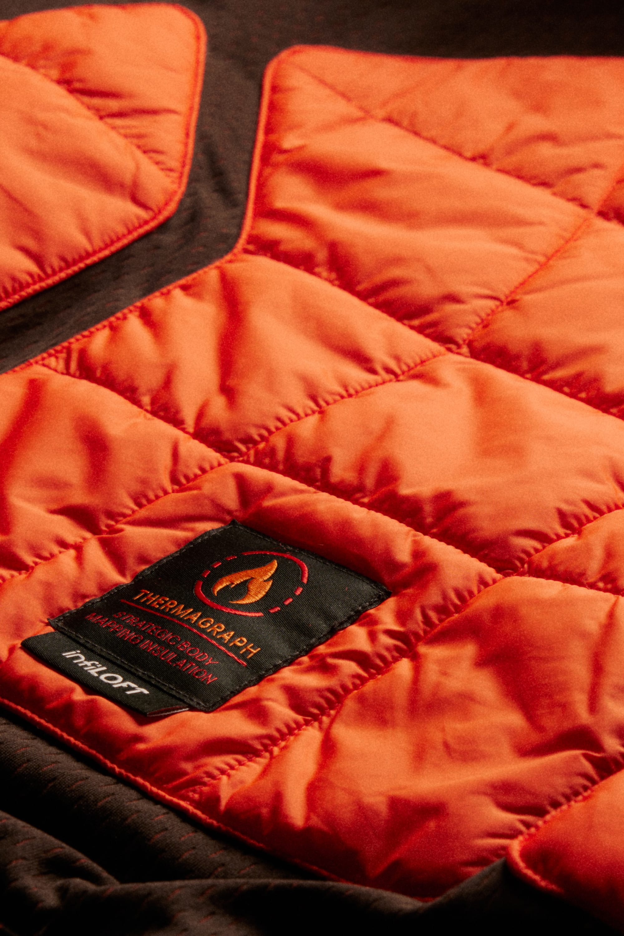 Close-up of an orange quilted jacket with a black Thermagraph® label featuring a flame icon and the word MIDFIT, laying on a dark brown fabric background, next to a pair of 686 Men’s Hydra™ Thermagraph® Pants by 686.