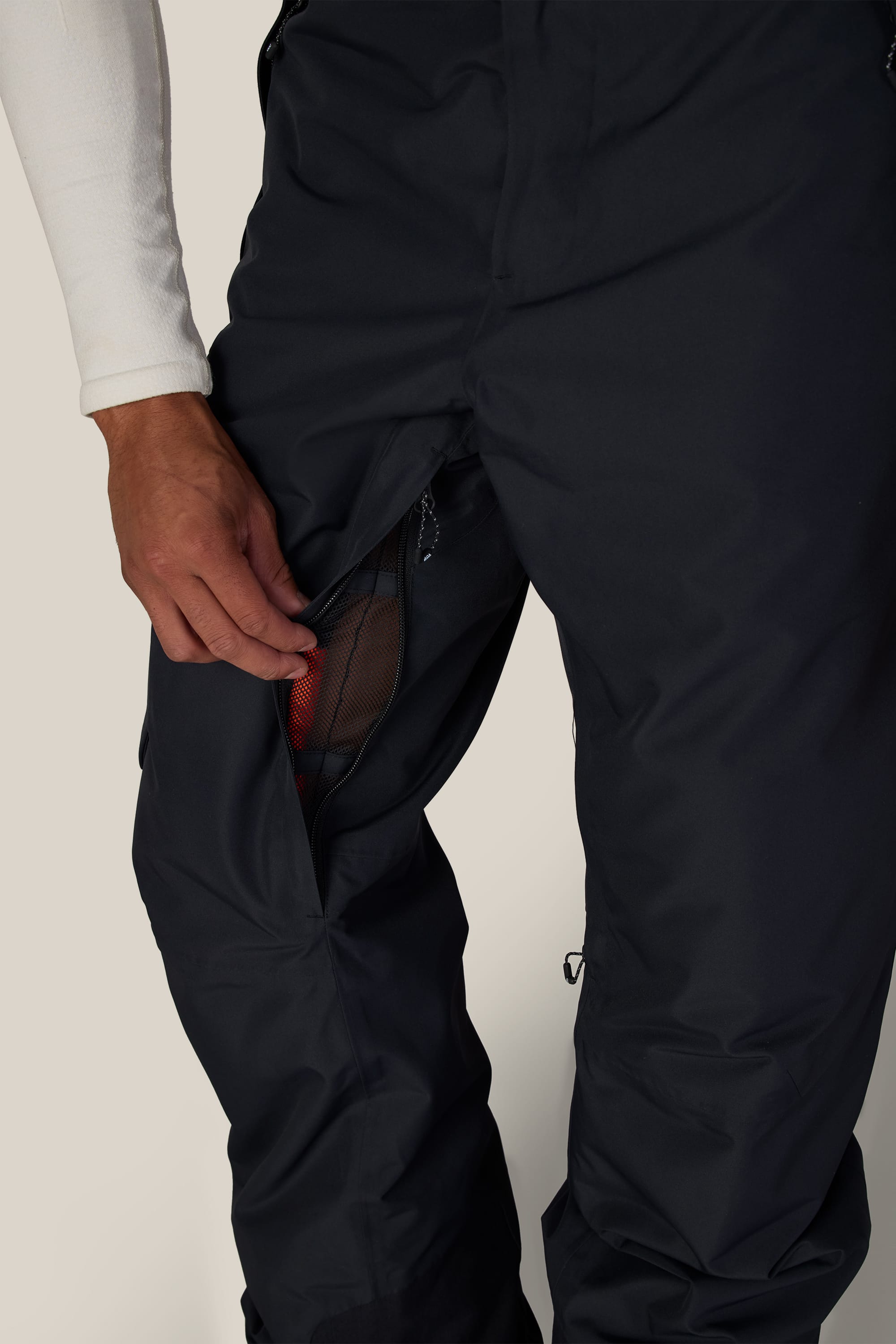 A person wearing the 686 Men’s Hydra™ Thermagraph® Pant in black unzips a mesh-lined thigh vent, revealing red fabric underneath. Their hand is pulling the zipper, and they are dressed in a white long-sleeve top.