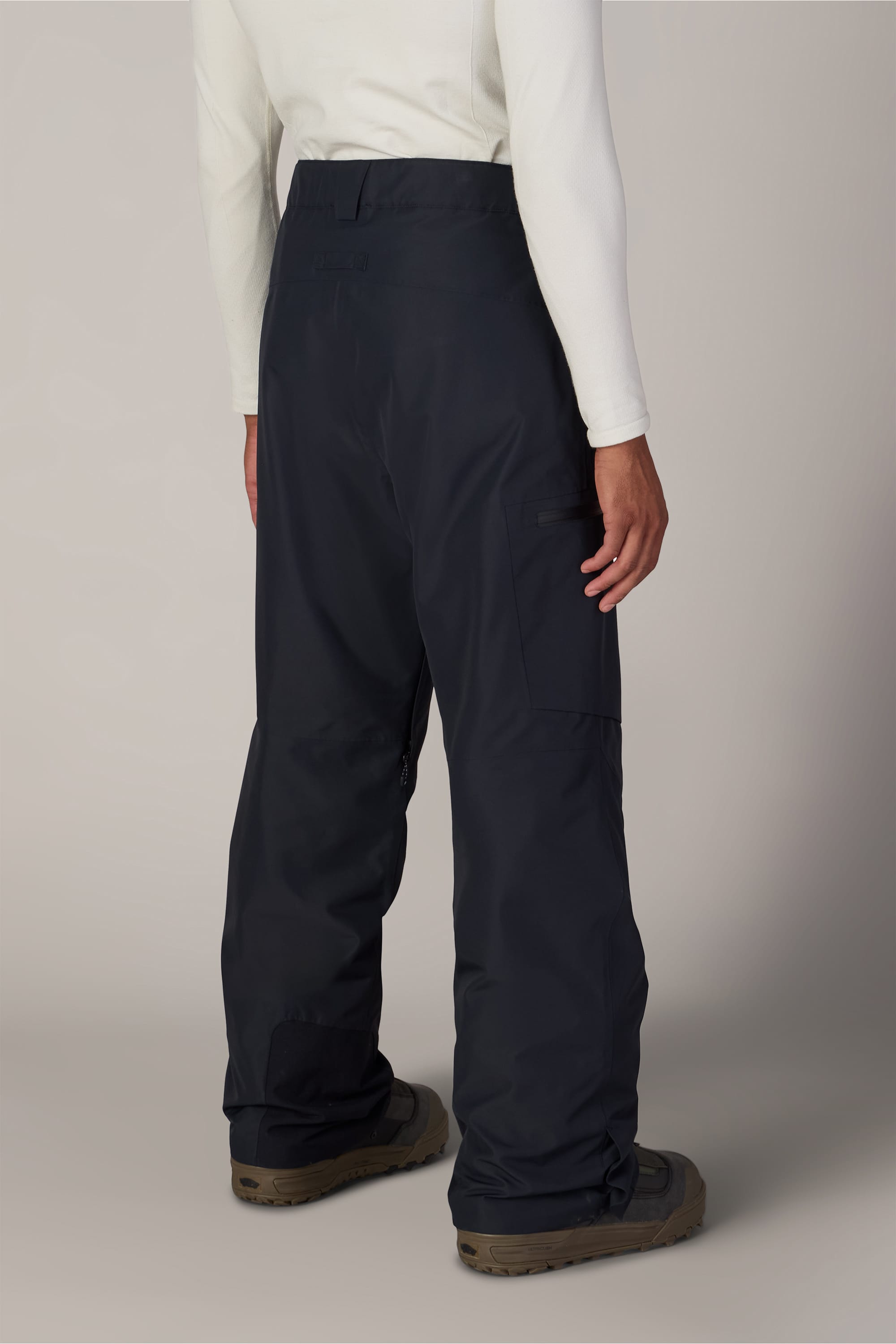 A person wearing a white long-sleeve shirt, 686 Men’s Hydra™ Thermagraph® Pant by 686 in navy blue, and brown shoes is standing facing away from the camera against a plain background.