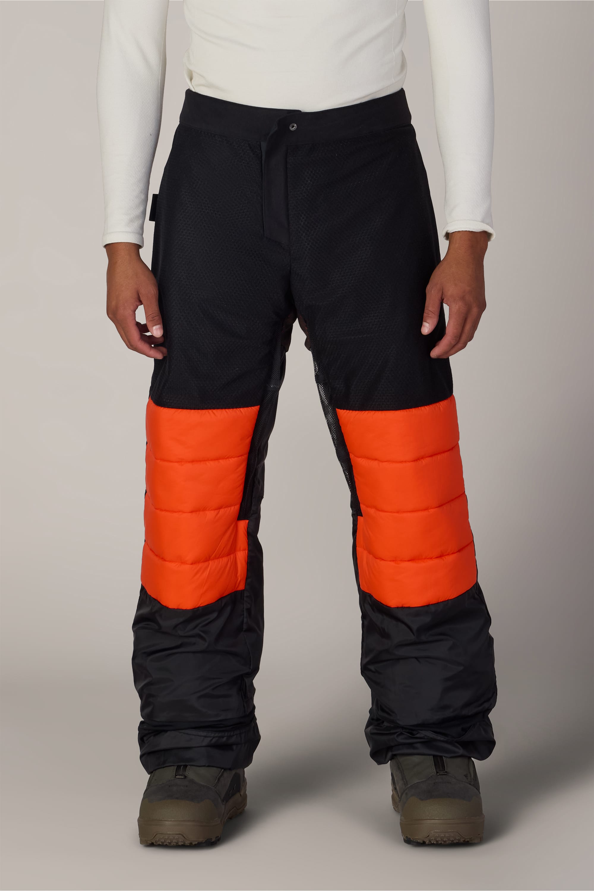 A person wearing 686 Men’s Hydra™ Thermagraph® Pants in black with bright orange padded sections on the thighs, paired with a white long-sleeve top and brown boots, standing against a plain background.