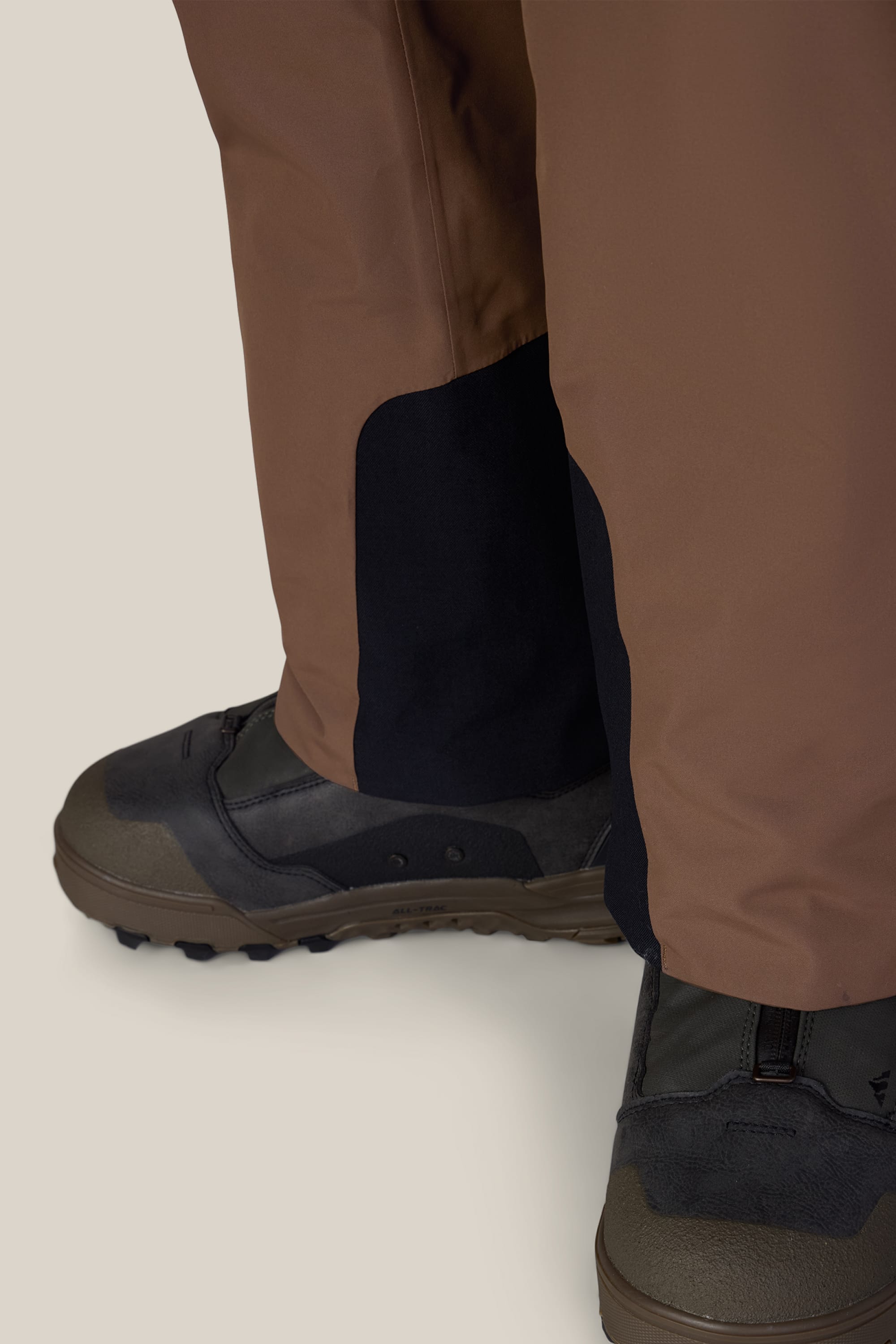 Close-up of 686 Men’s Hydra™ Thermagraph® Pants in brown, featuring black reinforced ankle patches, worn over dark hiking shoes with rugged soles, shown against a light background.