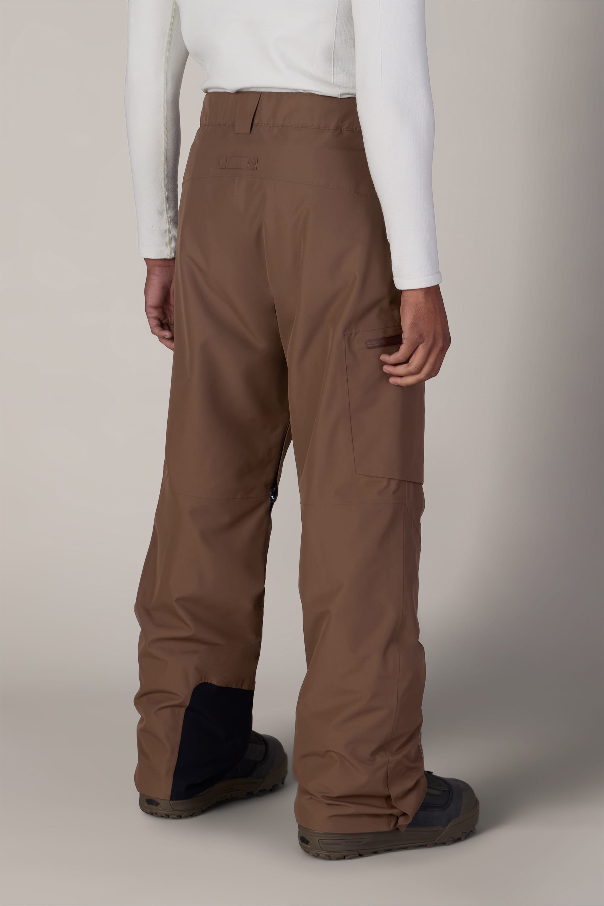 A person is shown from the back against a plain background, wearing the 686 Men’s Hydra™ Thermagraph® Pant in brown, paired with a white long-sleeve shirt and dark boots.