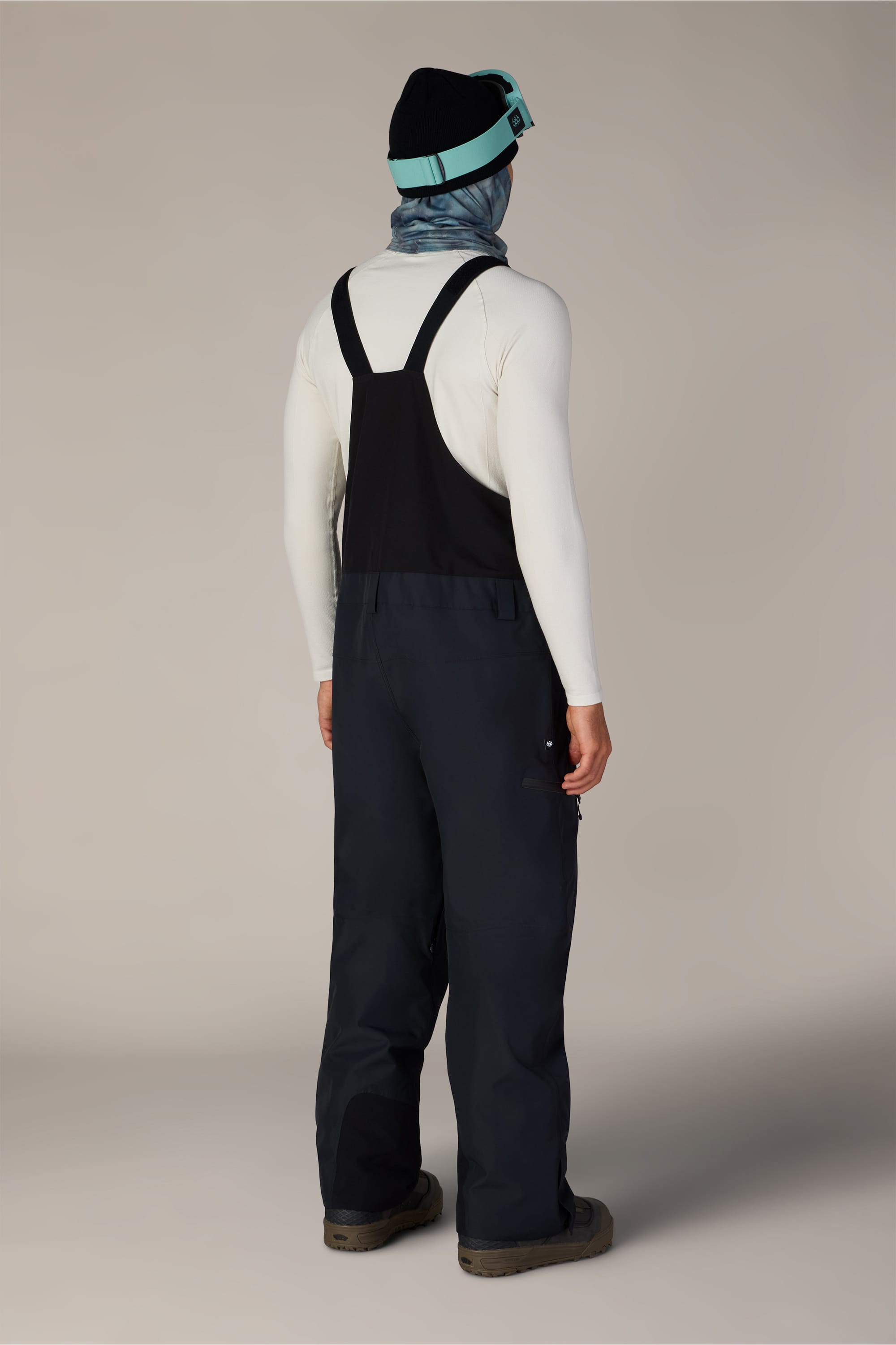 A person stands facing away against a plain background, wearing a black beanie, blue goggles, a blue neck gaiter, a white long-sleeve shirt, brown boots, and the 686 Men’s Lockup™ Shell Bib by 686.