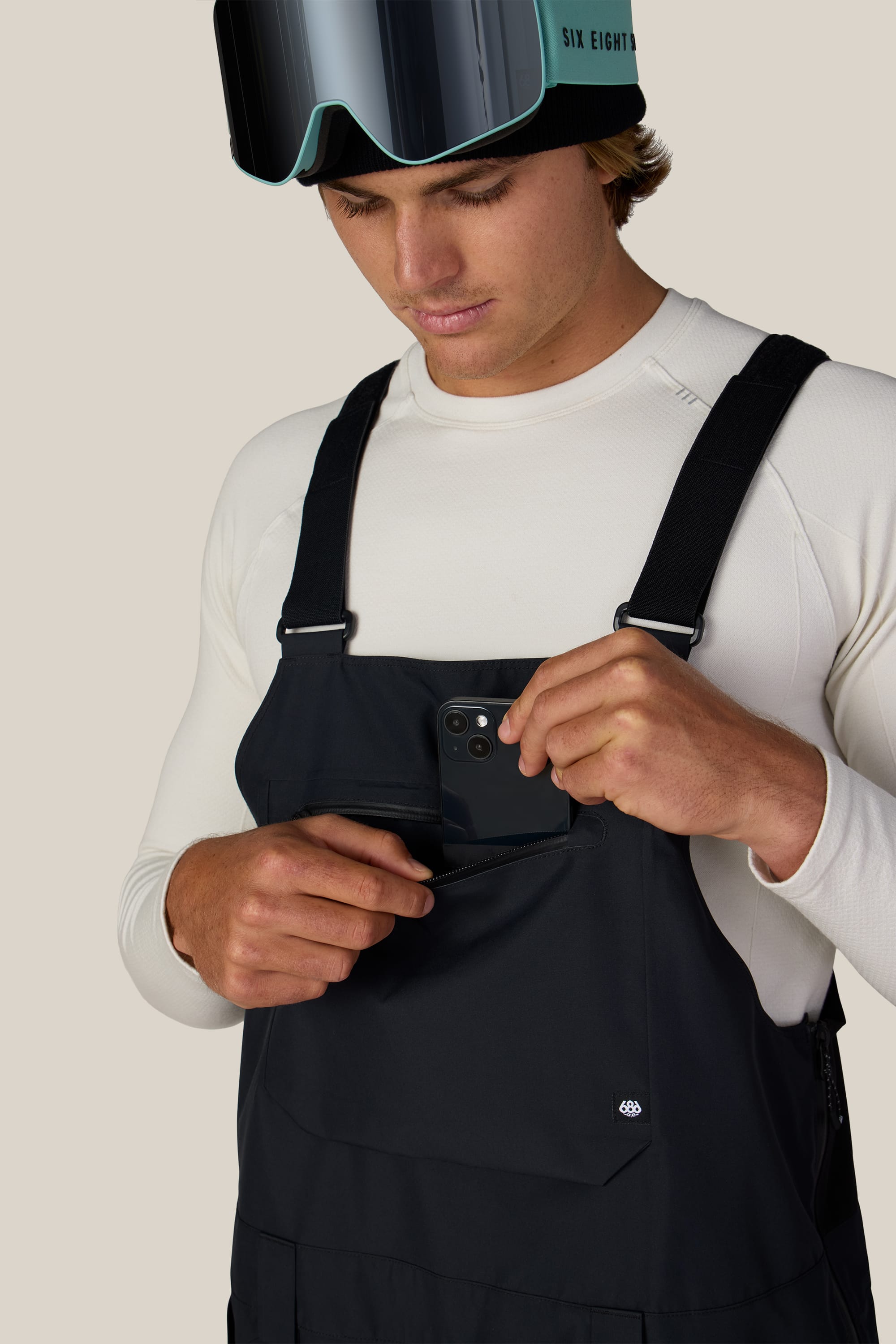 Wearing a white shirt and ski goggles, a person puts a phone into the chest pocket of the black 686 Men’s Lockup™ Shell Bib by 686, looking down at the pocket.