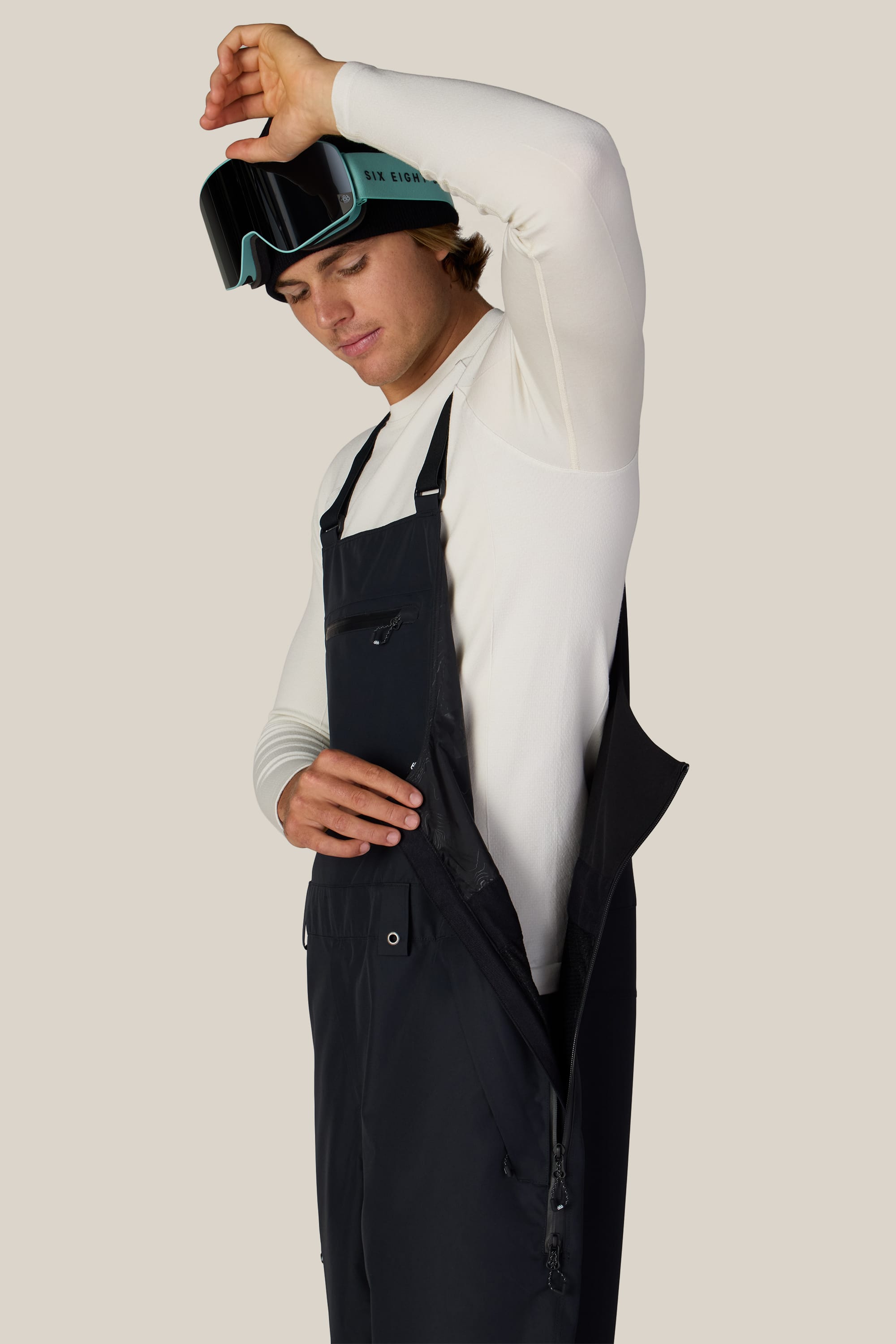 A person dressed in a white long sleeve shirt unzips the side of the 686 Men’s Lockup™ Shell Bib by 686. They have ski goggles resting on their forehead and are standing against a plain beige background.