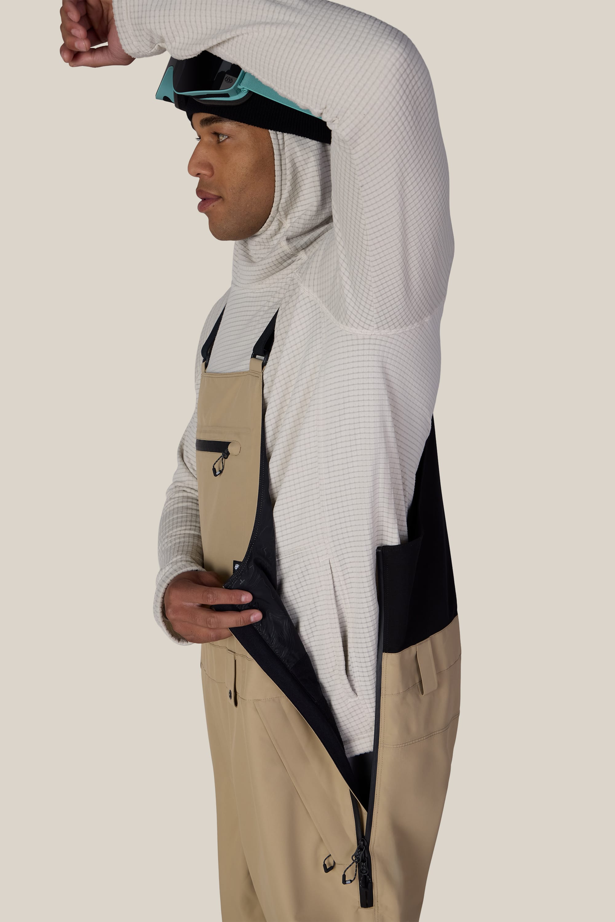 Wearing the 686 Men’s Lockup™ Shell Bib by 686, a person in a white hoodie, beige overalls, and ski goggles unzips the side ventilation panel to showcase the bib’s breathability and functionality.