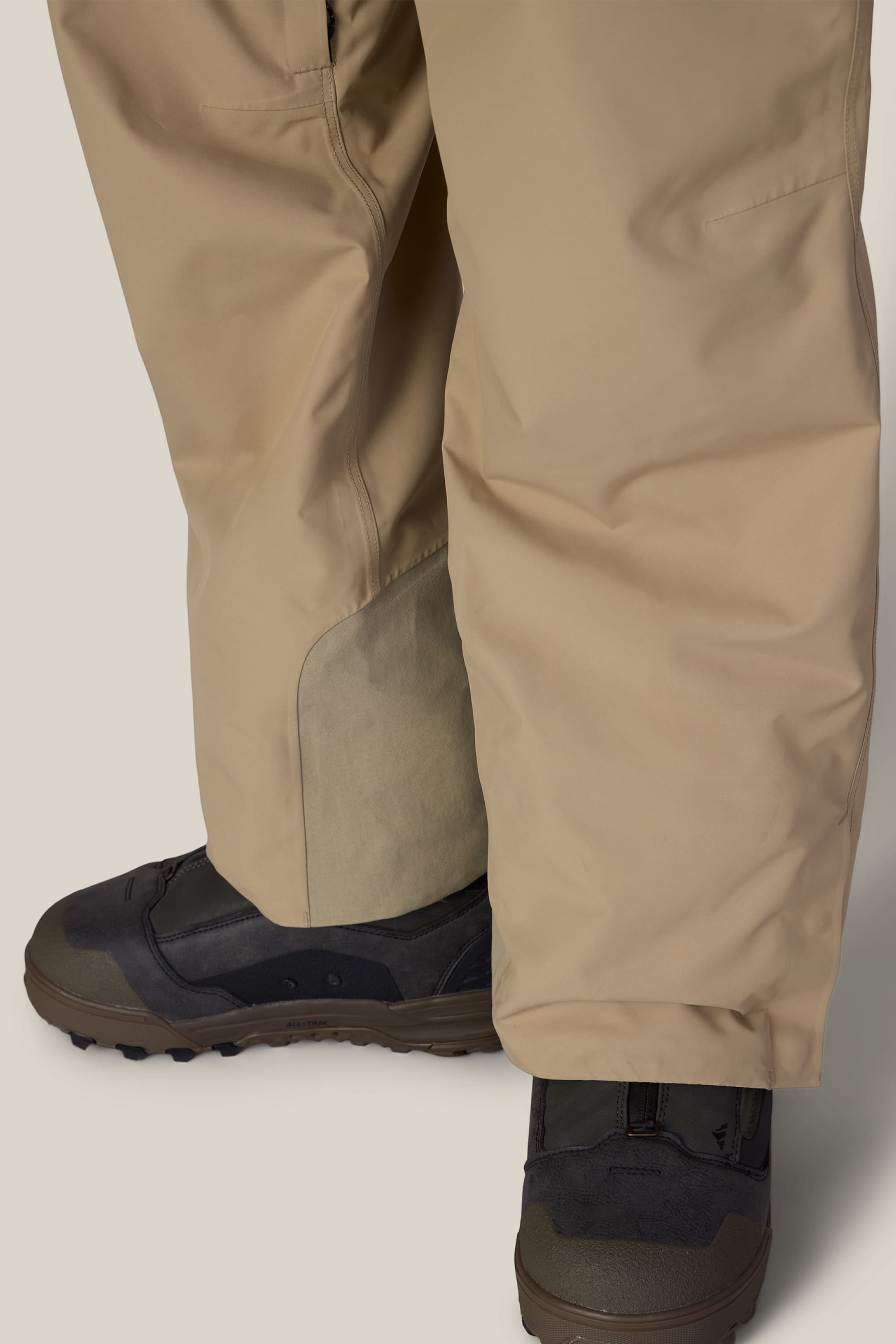 Close-up of a person wearing the 686 Men’s Lockup™ Shell Bib in tan, layered over dark brown and black hiking or snow boots, standing on a light background.