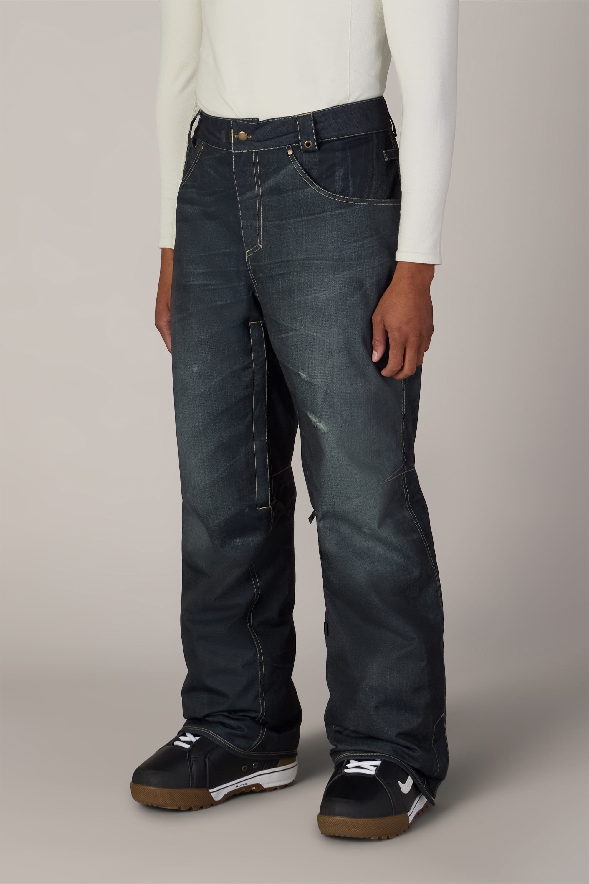 A person wearing a long-sleeve white shirt, the 686 Men’s Deconstructed™ Denim Insulated Pant by 686—loose dark blue woven pants with yellow stitching—and black and white sneakers, standing against a plain light gray background.