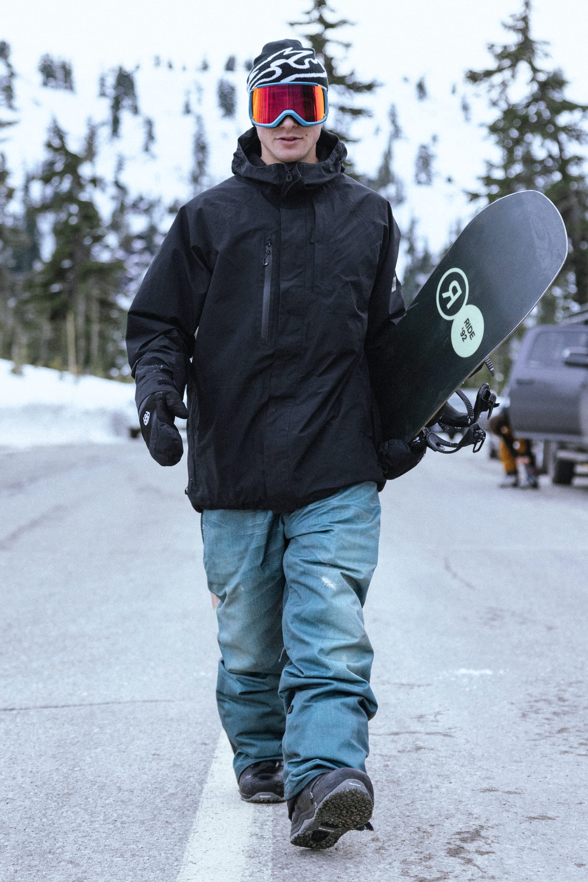 A person wearing a black jacket, 686 Men’s Deconstructed™ Denim Insulated Pant by 686, and goggles walks on a snowy road holding a snowboard. Snow and evergreen trees are visible in the background.