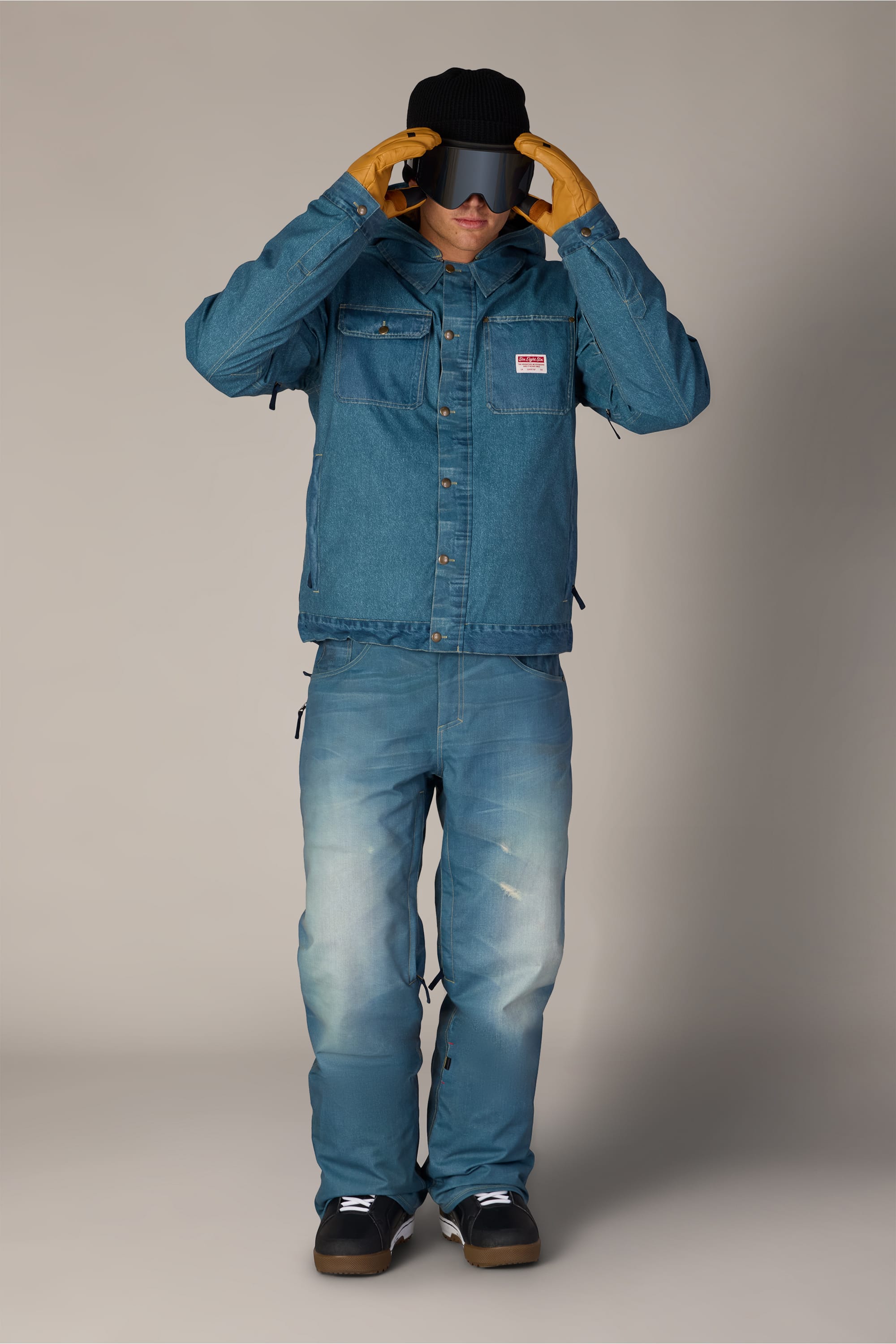 A person in a denim jacket and the 686 Men’s Deconstructed™ Denim Insulated Pant, wearing orange gloves, a black cap, and sunglasses, stands against a plain background and looks downward while adjusting the brim of their cap.