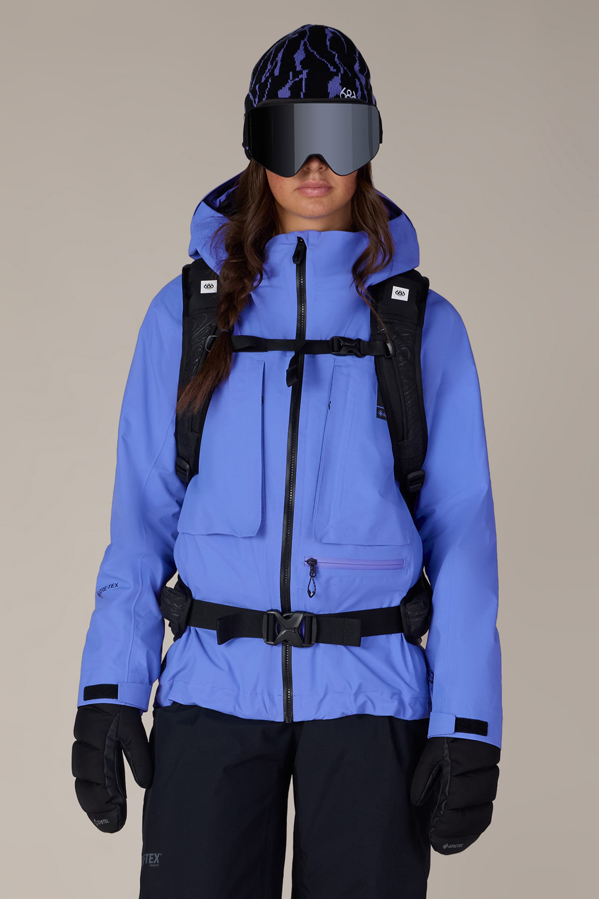 A person is standing facing forward, wearing the 686 Women’s GORE-TEX® Scheme Thermagraph® Jacket in bright blue with a hood, black snow pants, gloves, a black printed cap, large ski goggles, a backpack, and a black belt.