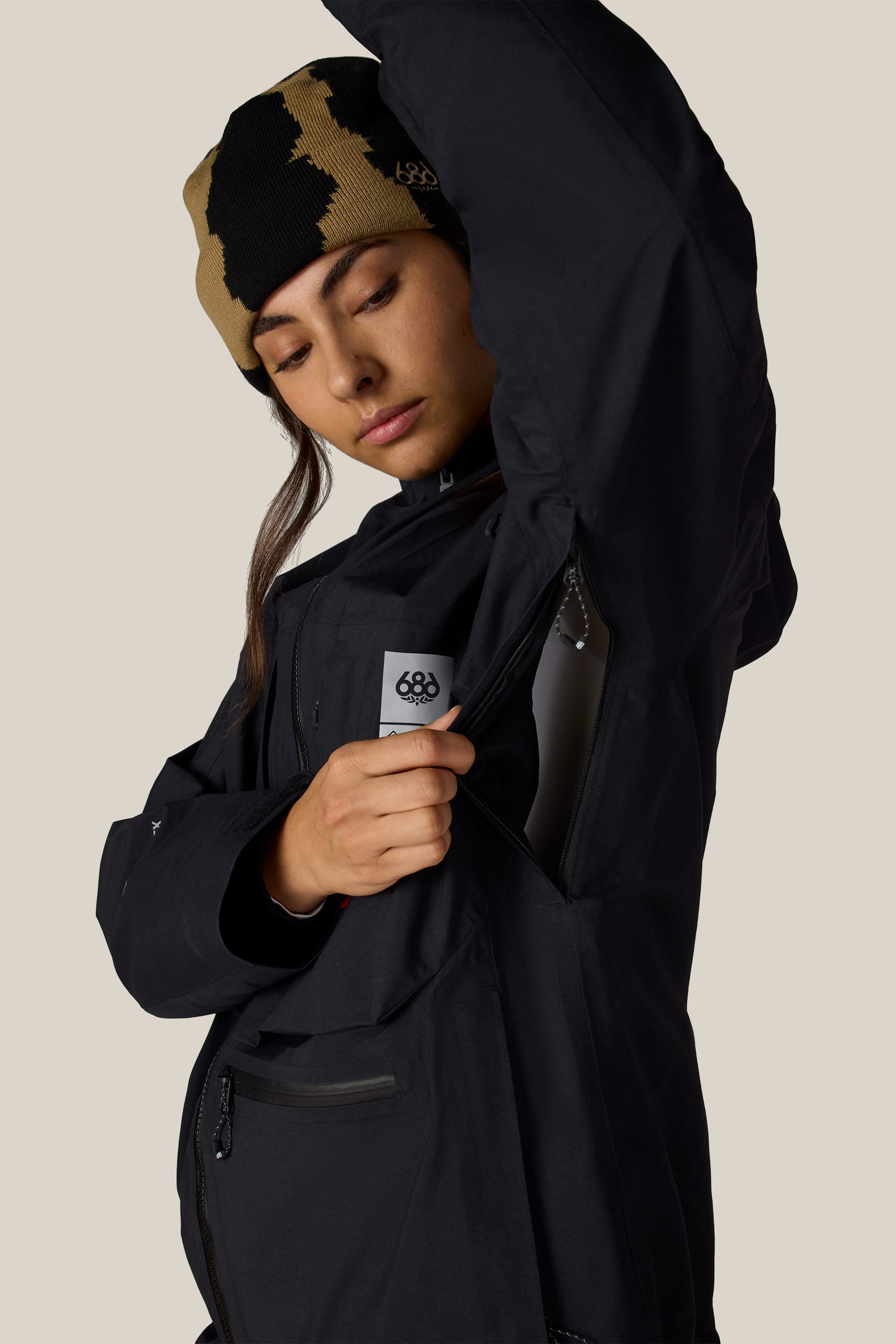 A person wearing the 686 Women’s GORE-TEX® Scheme Thermagraph® Jacket in black and a tan-and-black beanie unzips a vent under their arm, revealing the jacket’s mesh lining. The background is plain and light-colored.