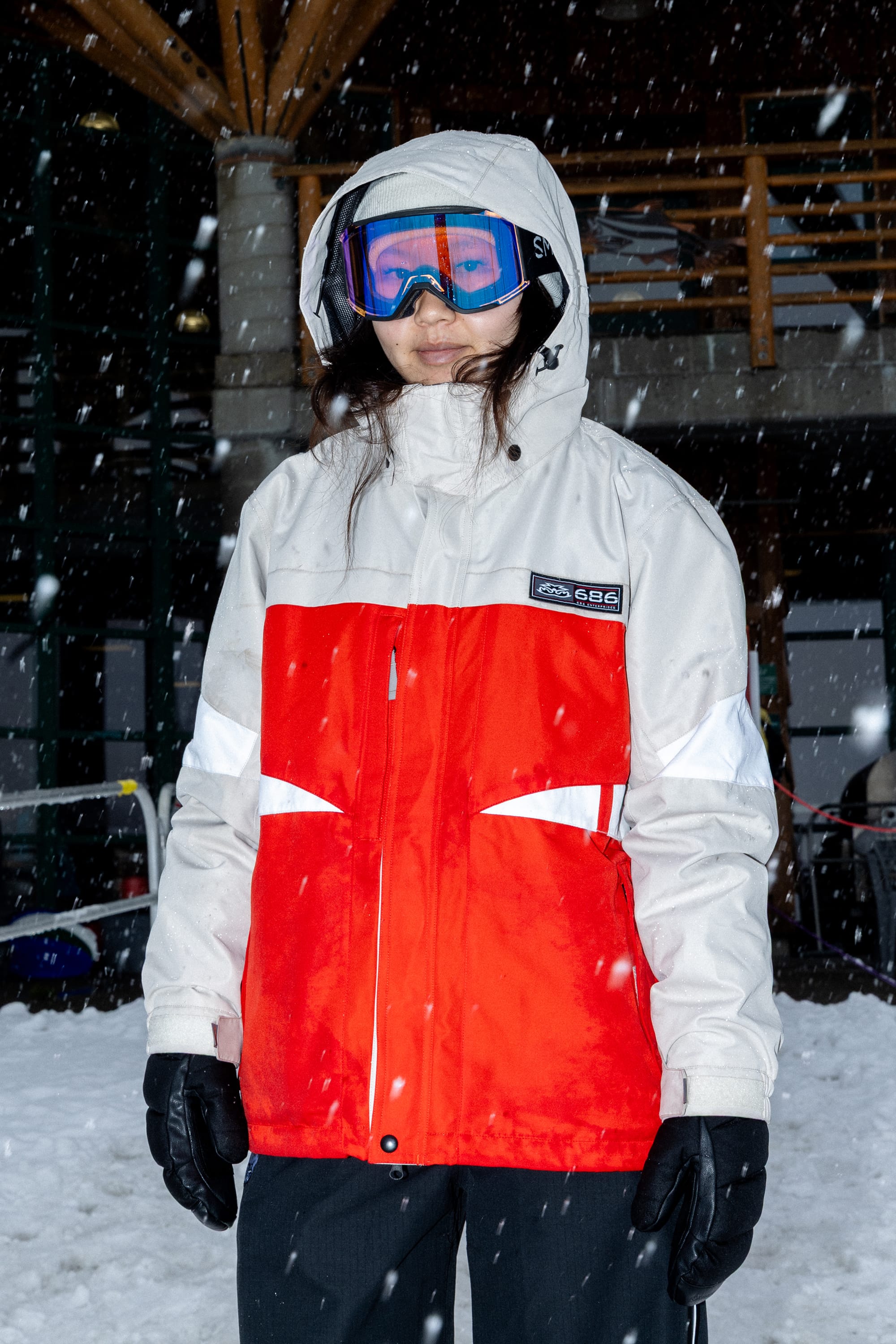 A person wearing the 686 Women’s Search Jacket in red and white, along with black gloves and blue ski goggles, stands outside in falling snow with a snowy building and wooden railing in the background.