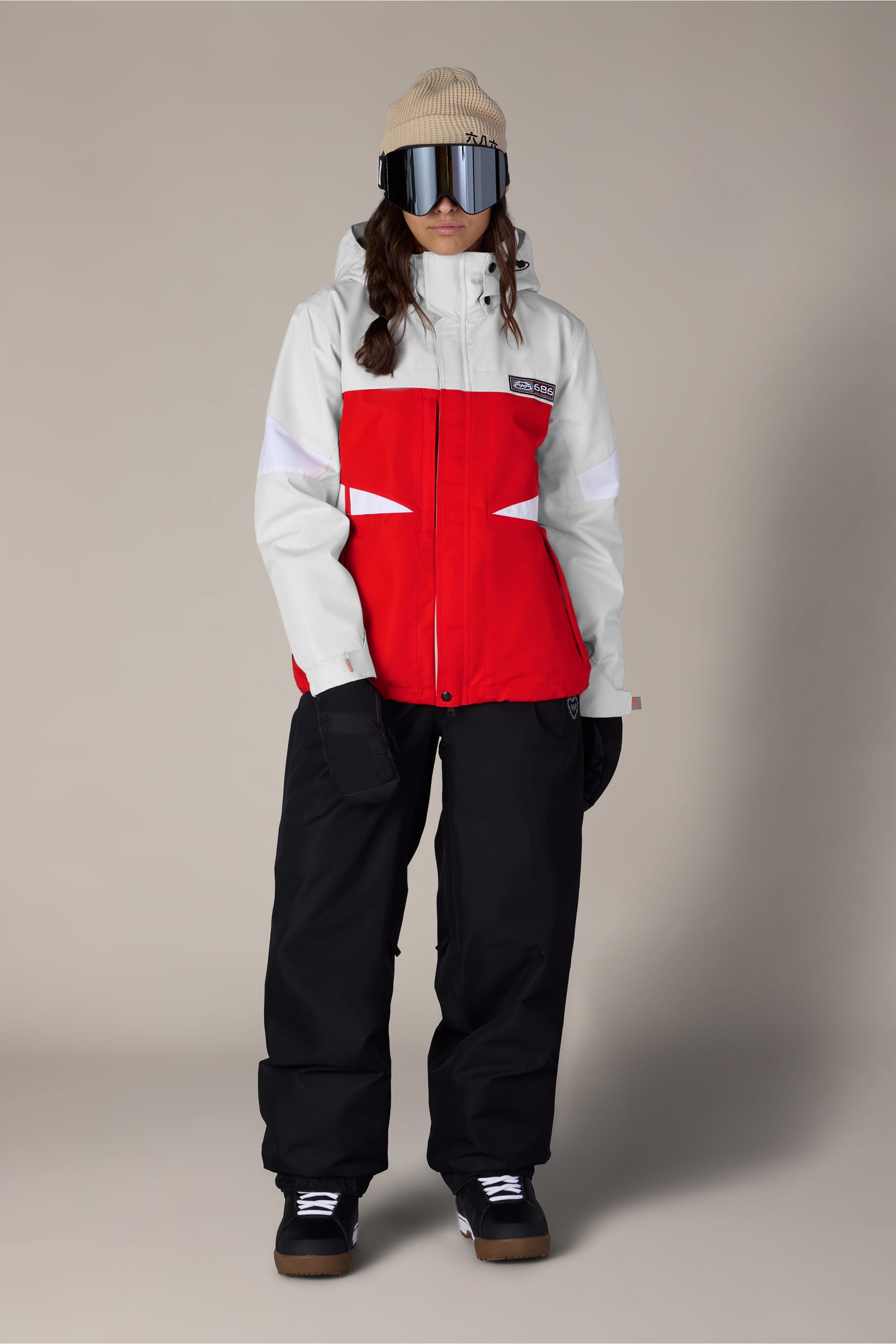 A person wearing the 686 Women’s Search Jacket in red and white, paired with black snow pants, black gloves, a beige beanie, and reflective ski goggles stands facing forward against a plain background.