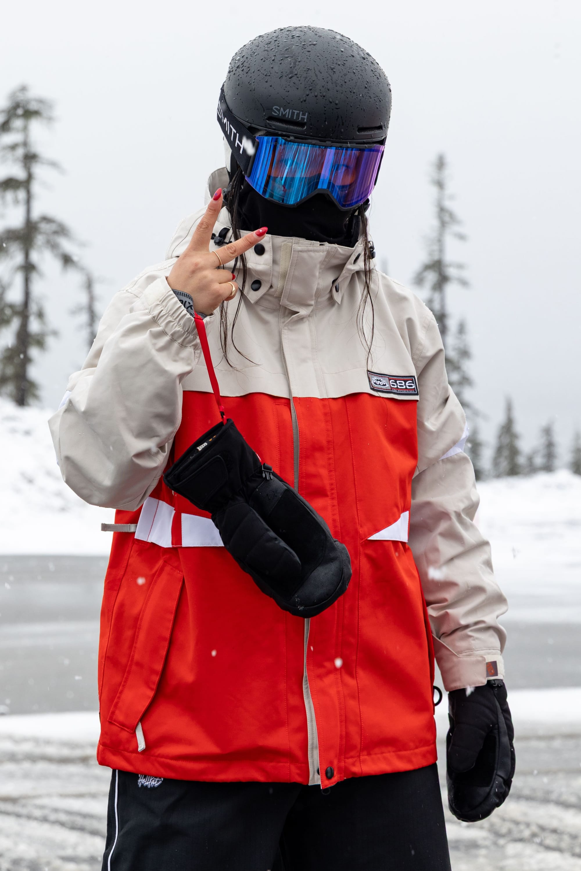 Wearing the 686 Women’s Search Jacket by 686 in red and beige, along with a black helmet and goggles, a person stands in a snowy landscape, flashing a peace sign with one hand while holding black mittens in the other.