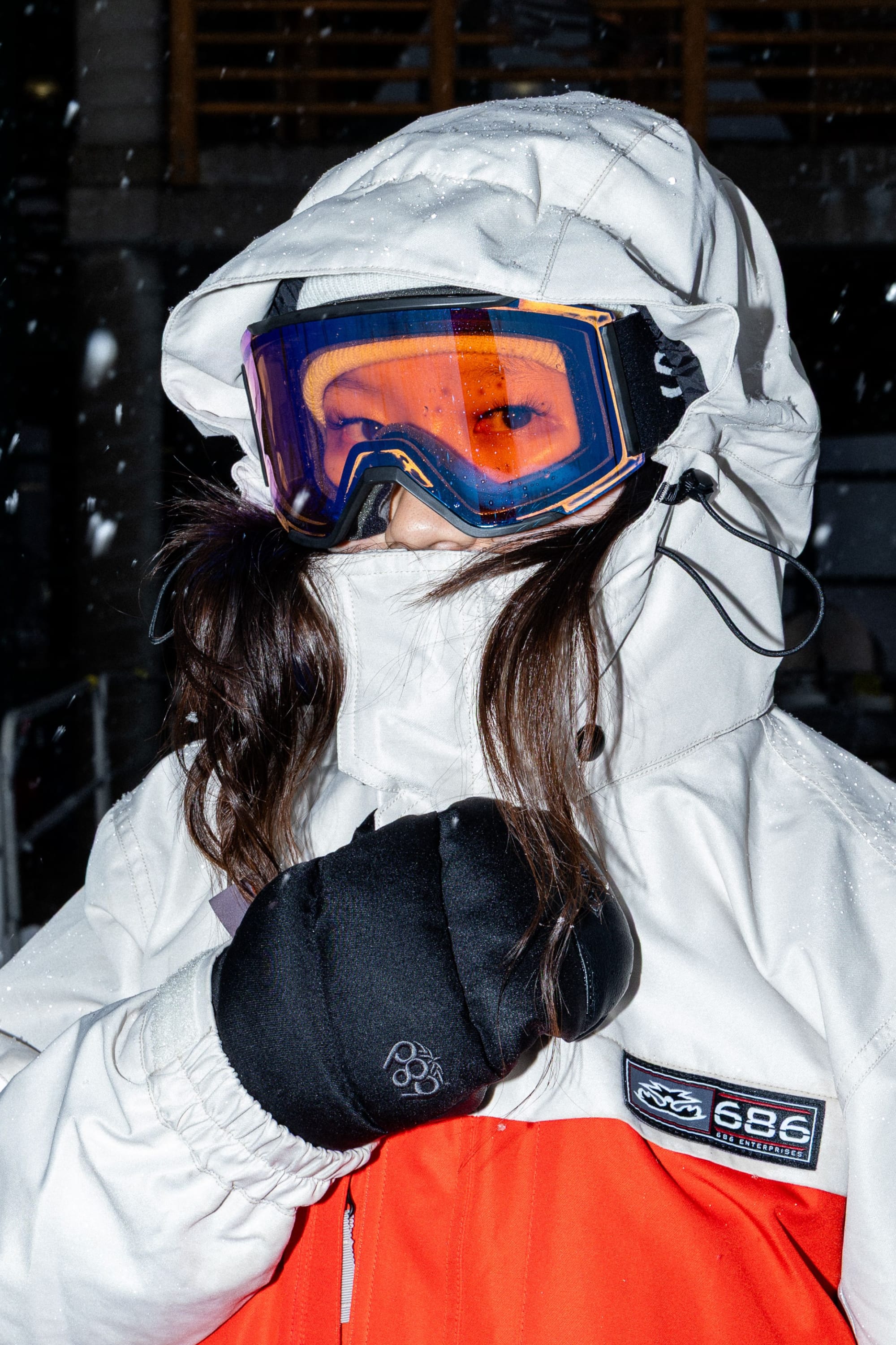 Wearing the 686 Women’s Search Jacket by 686 along with ski goggles and black gloves, a person stands outside in falling snow, looking at the camera.