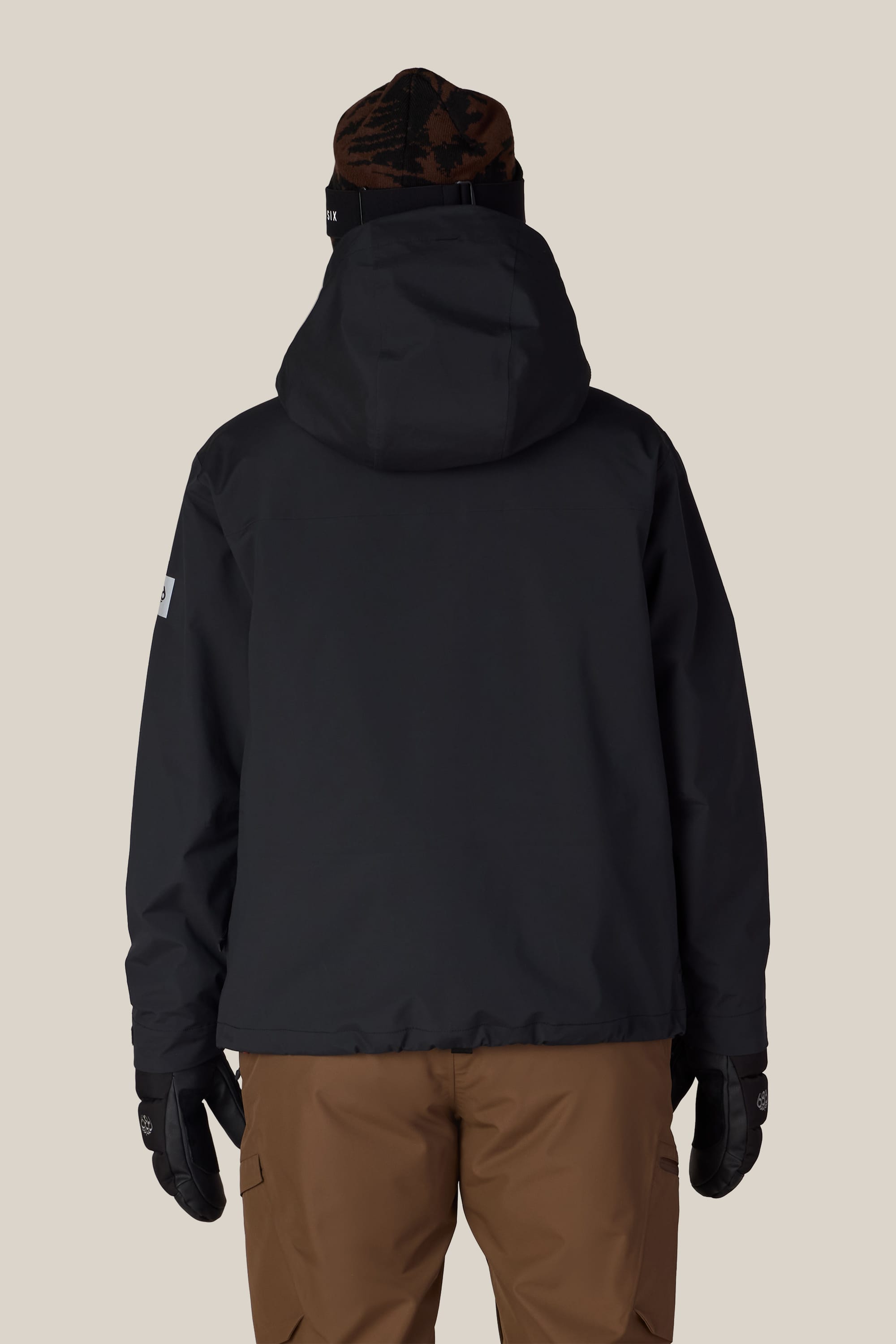 A person is shown from the back, standing against a plain beige background, wearing the 686 Women’s SMARTY® 3-in-1 Framework™ 3L Jacket, along with brown pants, black gloves, and a beanie.