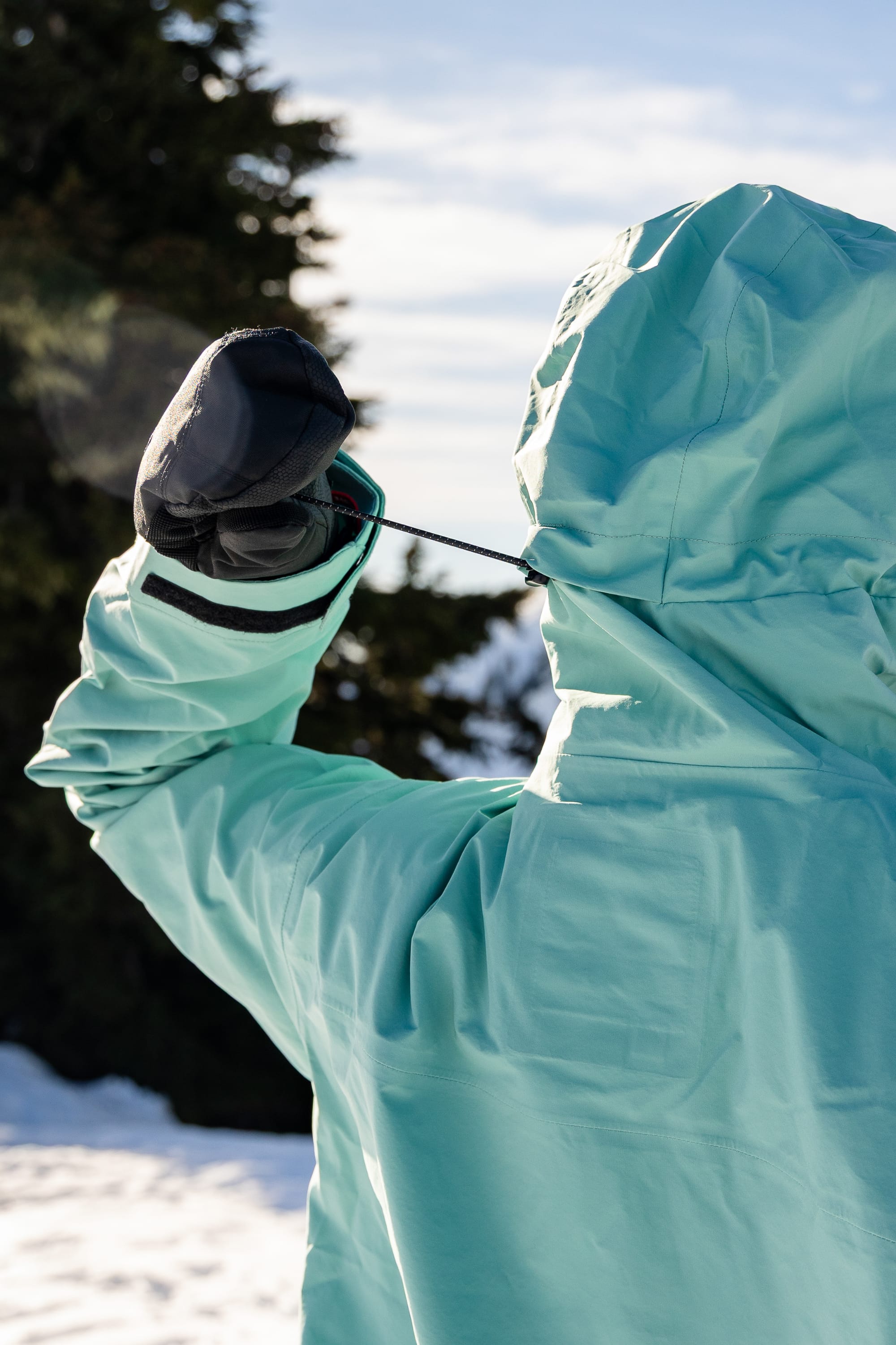 Wearing the 686 Women’s SMARTY® 3-in-1 Framework™ 3L Jacket in light blue, a person adjusts their hood drawstring outdoors amidst snow and trees, pairing the jacket with black gloves.