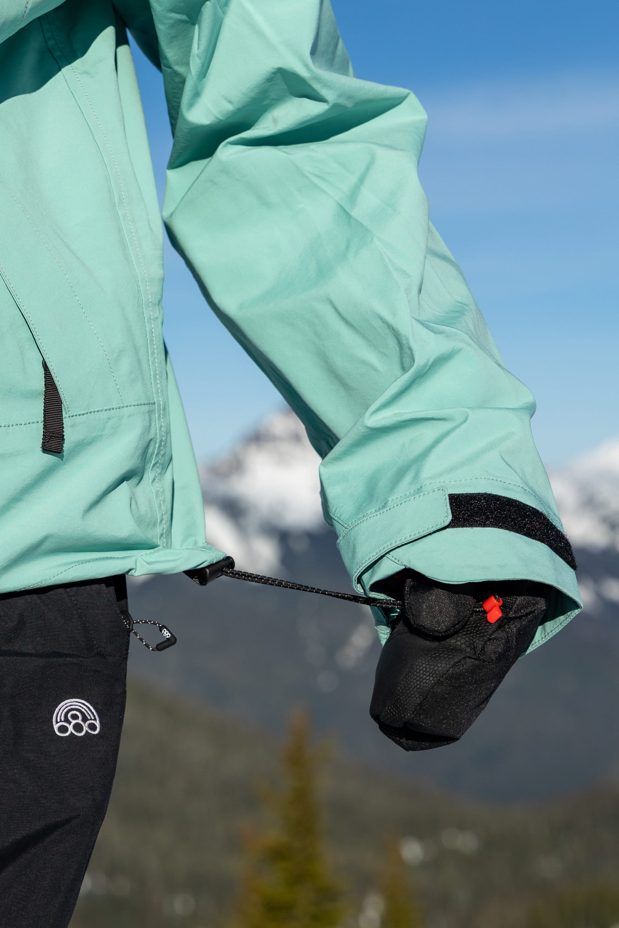 A person wearing the 686 Women’s SMARTY® 3-in-1 Framework™ 3L Jacket in teal and black gloves tightens a drawstring at the hem, with mountains and trees blurred in the background.