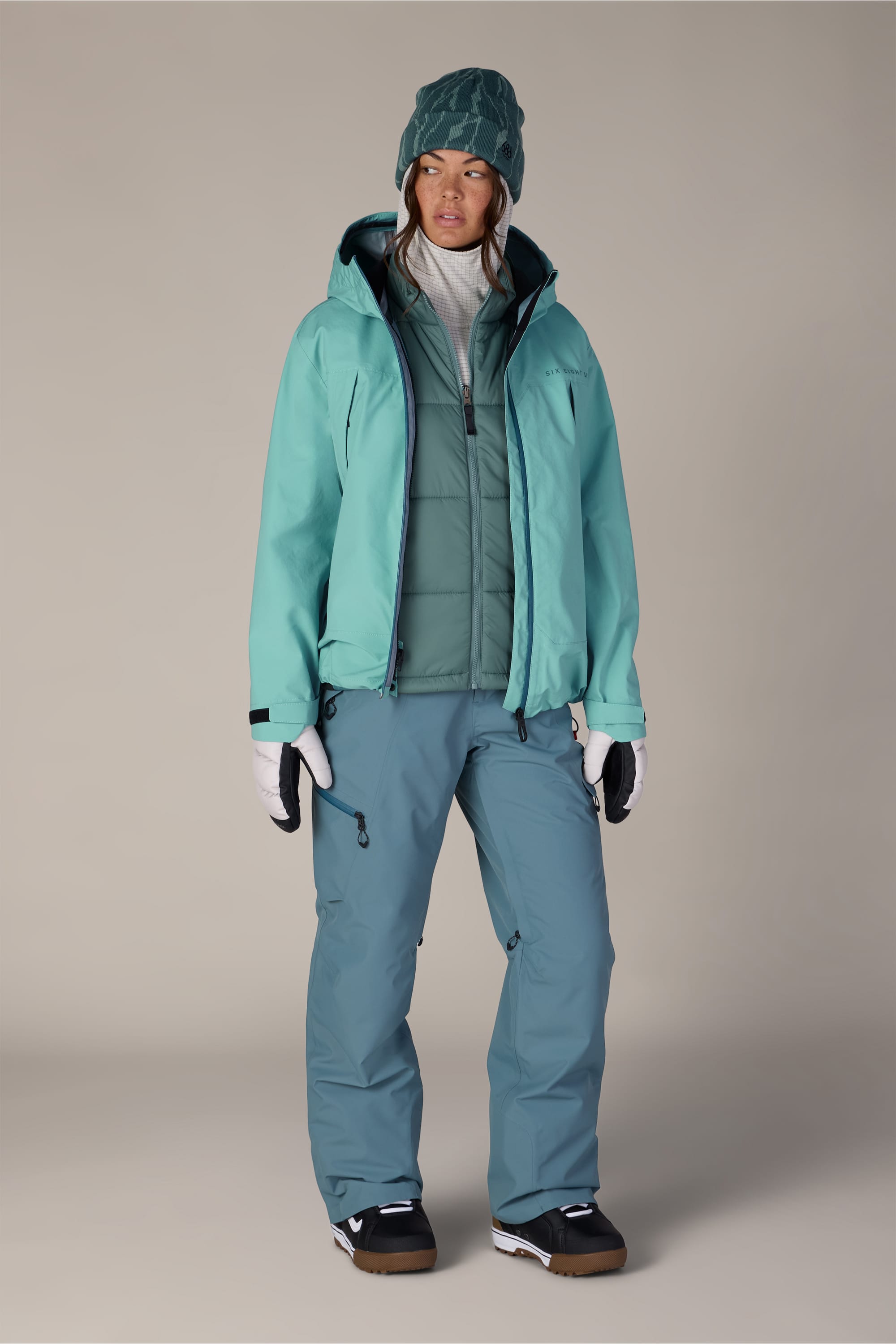 A person wearing the 686 Women’s SMARTY® 3-in-1 Framework™ 3L Jacket in teal, paired with matching pants, white gloves, a green beanie, and black boots, standing against a plain background.