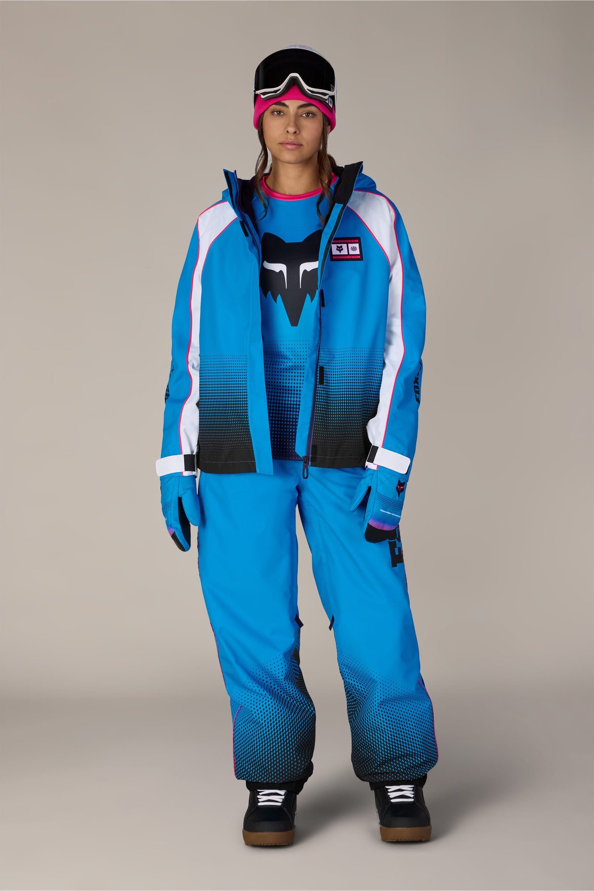 A person wearing a 686 Women’s Fox Racing 97 Shell Jacket, matching snow pants, a pink beanie, and goggles.