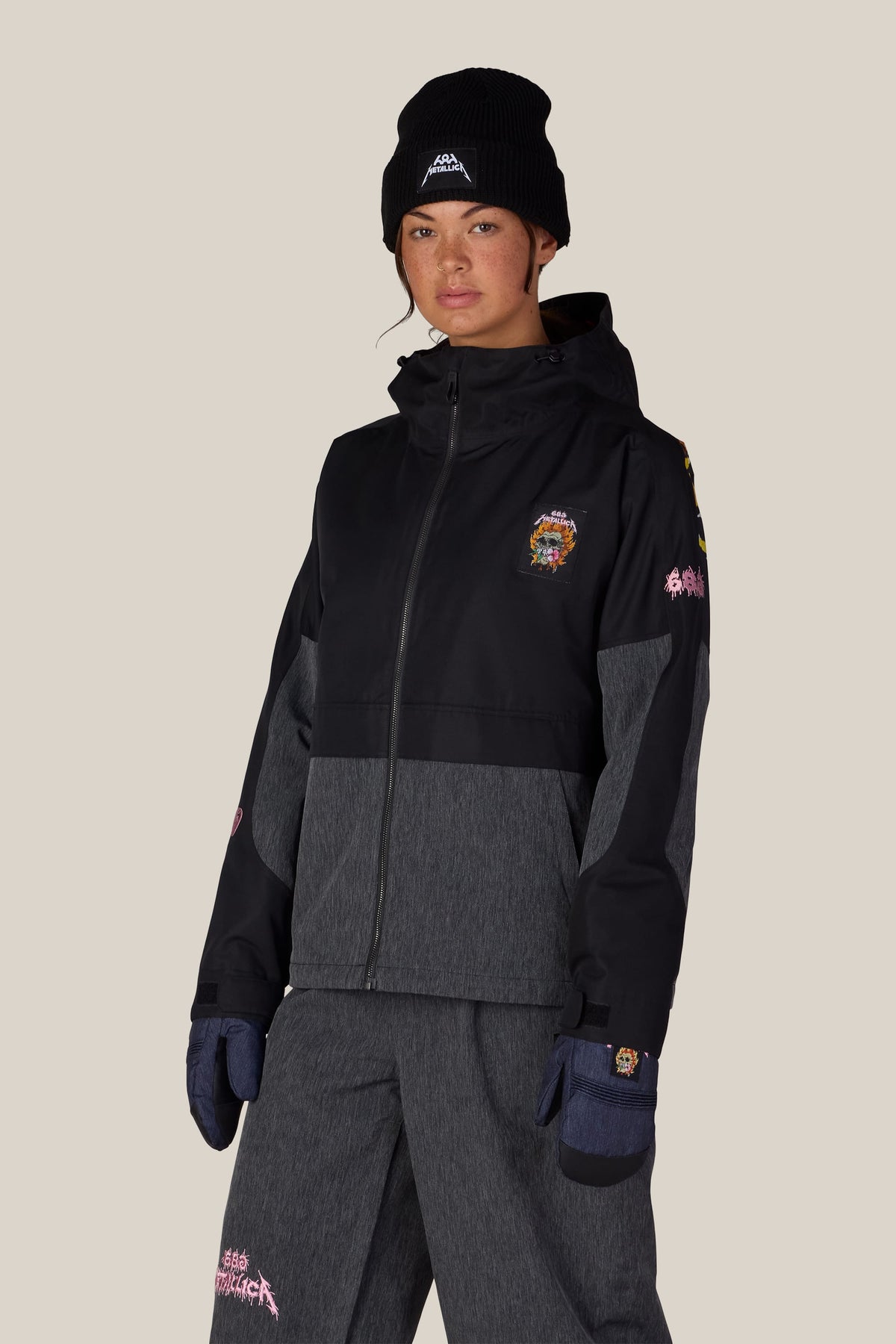A person wears a black beanie and a 686 Women’s Metallica Burning Skull Insulated Jacket featuring color-blocked dark gray panels, a hood, and skull and band-themed graphic patches.