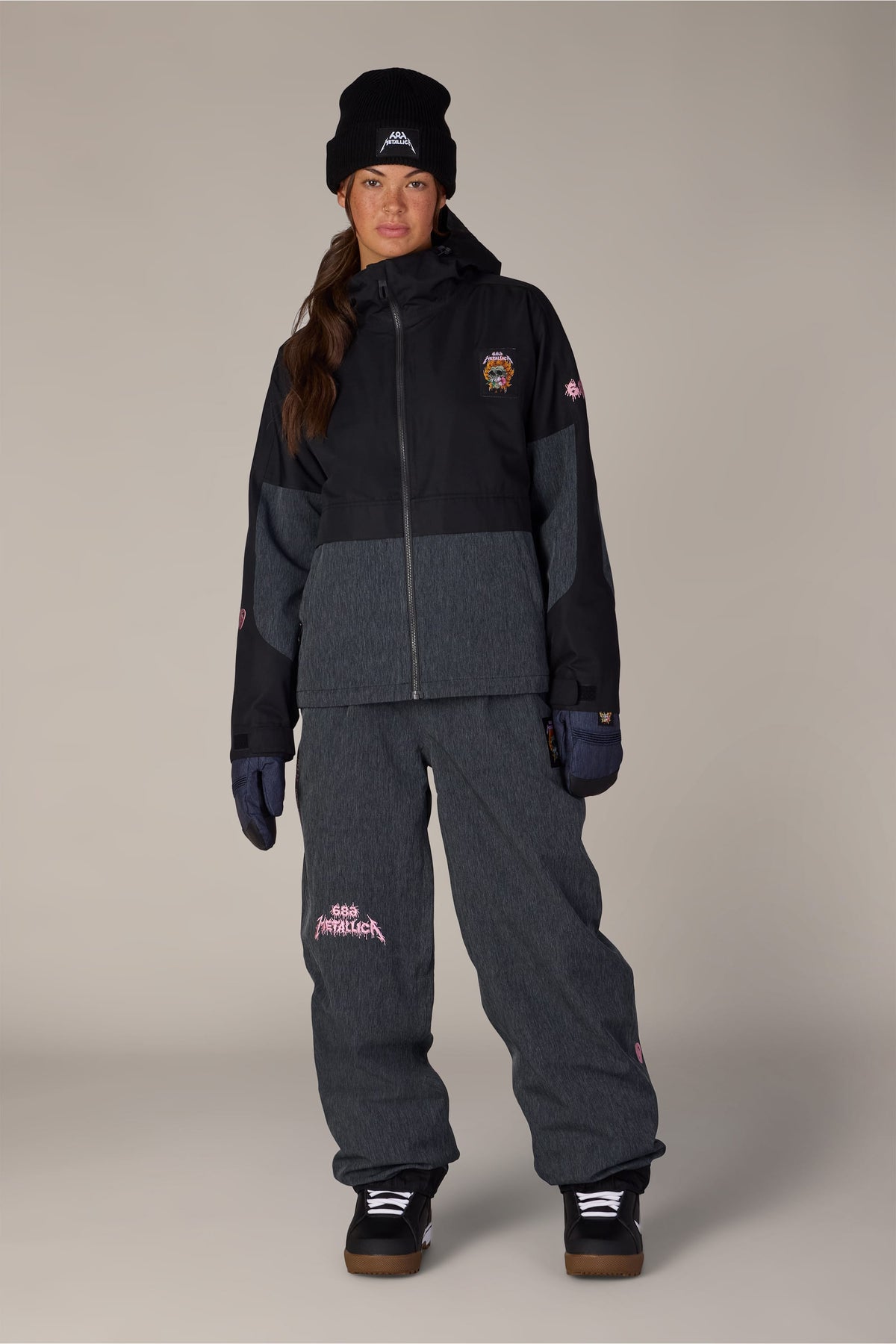 A person wearing a 686 Women’s Metallica Burning Skull Insulated Jacket and matching trousers, both decorated with Metallica skull graphics, paired with a black beanie, gloves, and boots.