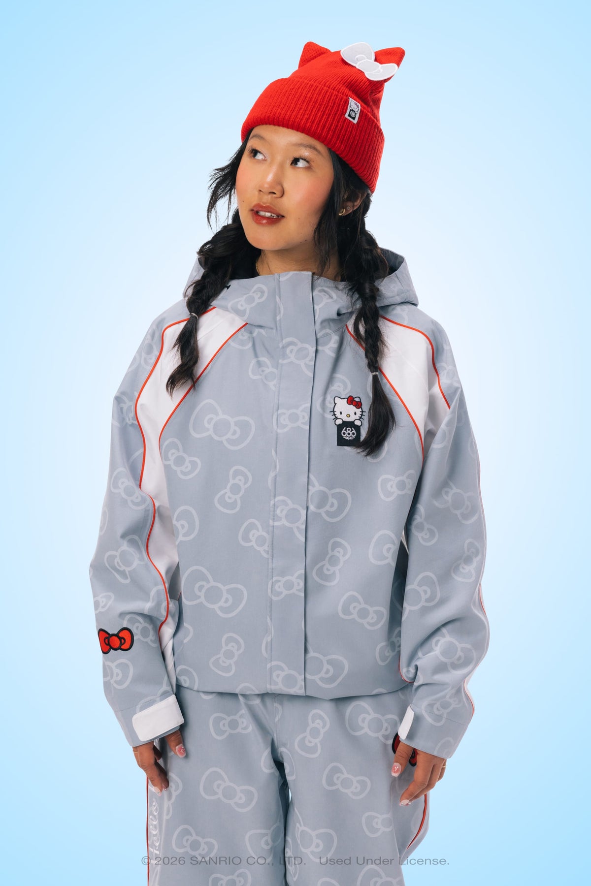 A woman stands against a light blue background, looking slightly upward, dressed in the 686 Women’s Hello Kitty® Lil Bow Shell Jacket with matching pants and a red beanie adorned with a bow. The outfit features light gray tones, red accents, and Hello Kitty graphics.