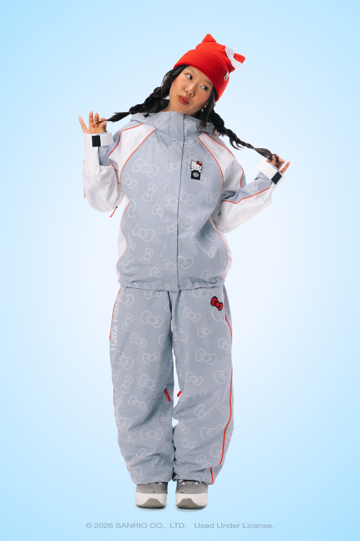 A person poses playfully, wearing the 686 Women’s Hello Kitty® Lil Bow Shell Jacket from 686 in light blue with red accents, gray sneakers, and a red beanie. The background is light blue.