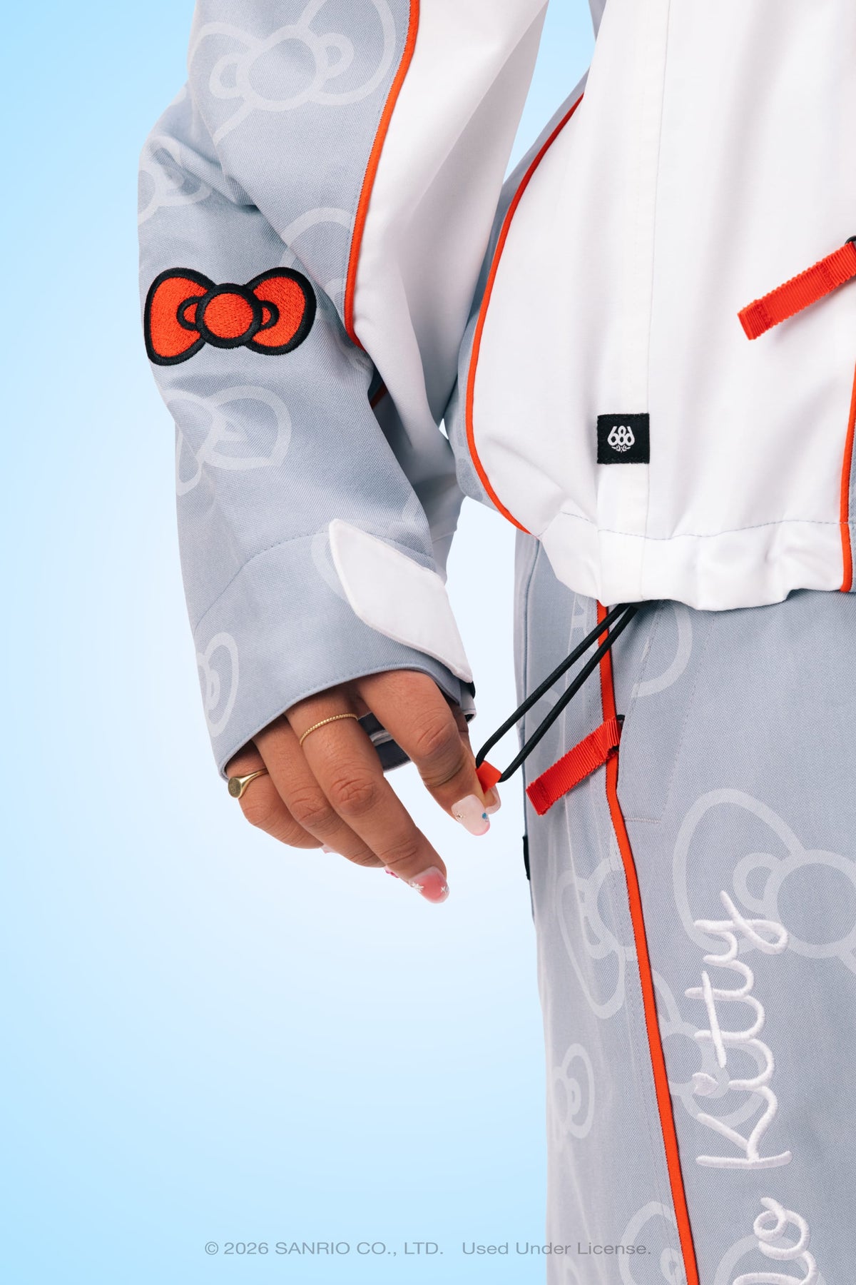 Wearing the 686 Women’s Hello Kitty® Lil Bow Shell Jacket, a person adjusts black drawstrings. The gray and white jacket from 686 features red accents and Hello Kitty® patterns, set against a light blue background.