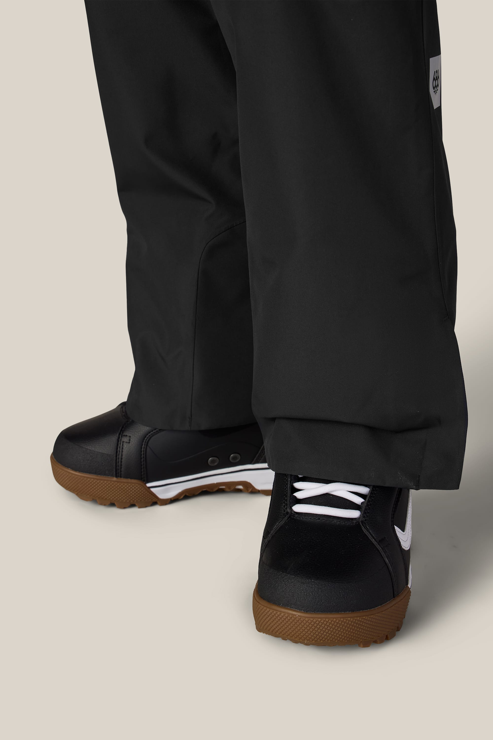 A close-up of a person wearing the 686 Women’s Geode Thermagraph® Pant, paired with black sneakers featuring white laces and brown soles, standing on a light beige background.