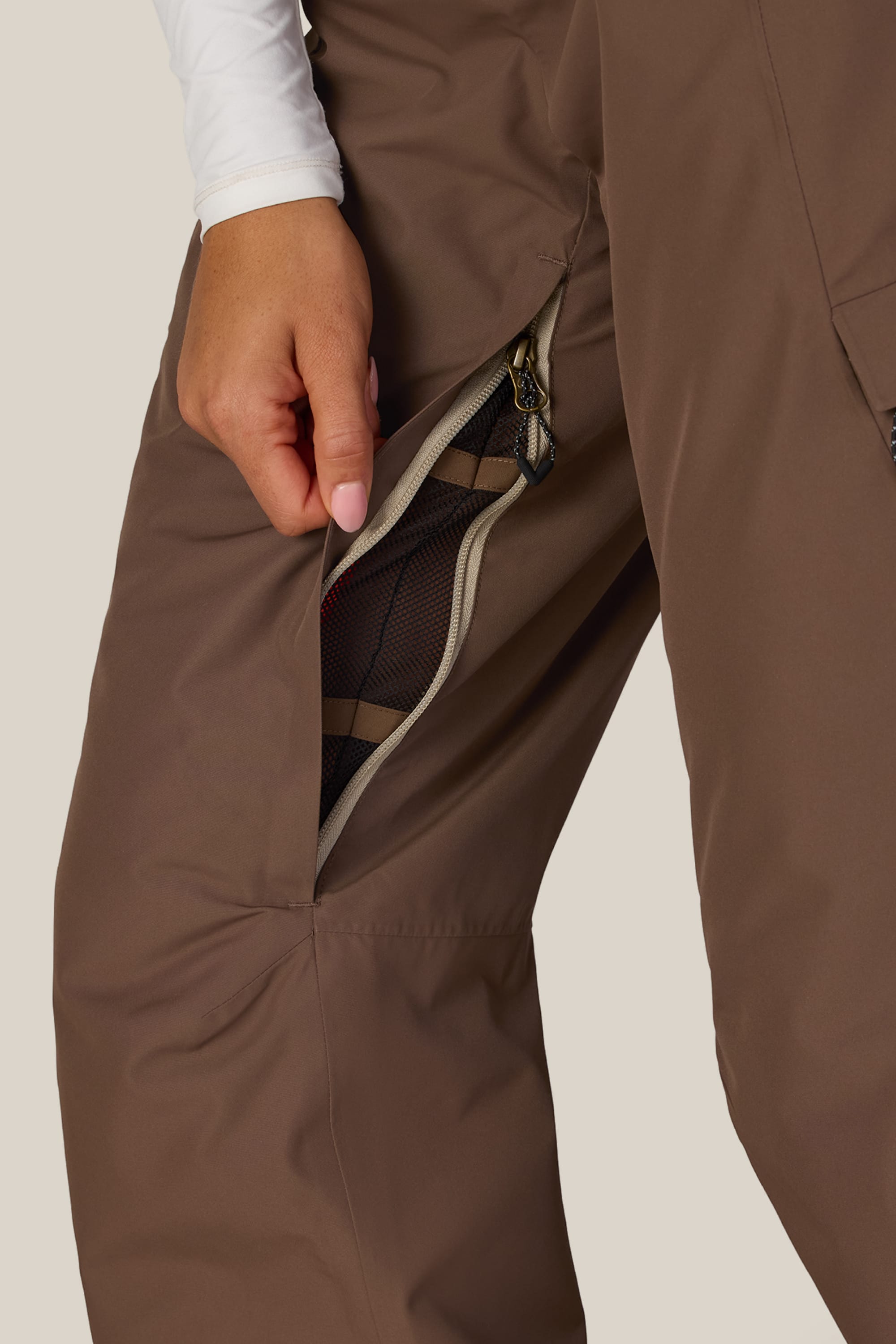 A person wearing 686 Women’s Geode Thermagraph® Pant in brown unzips a mesh-lined side vent, revealing the breathable inner layer. Only the lower torso and hand are visible against a neutral background.