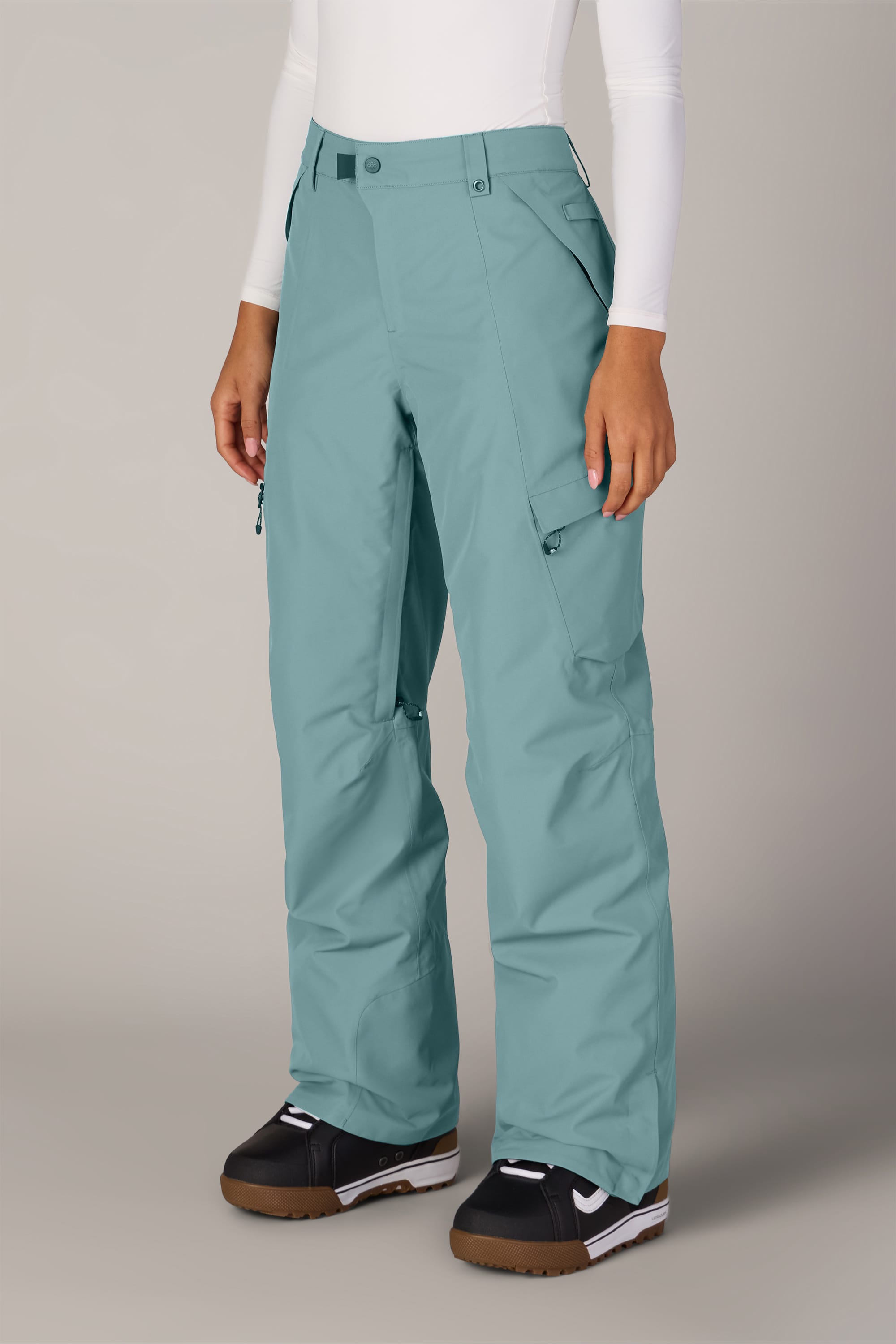 A person wearing teal 686 Women’s Geode Thermagraph® Pants with a relaxed fit and pockets, a white long-sleeve shirt, and black snow boots stands against a plain light background.