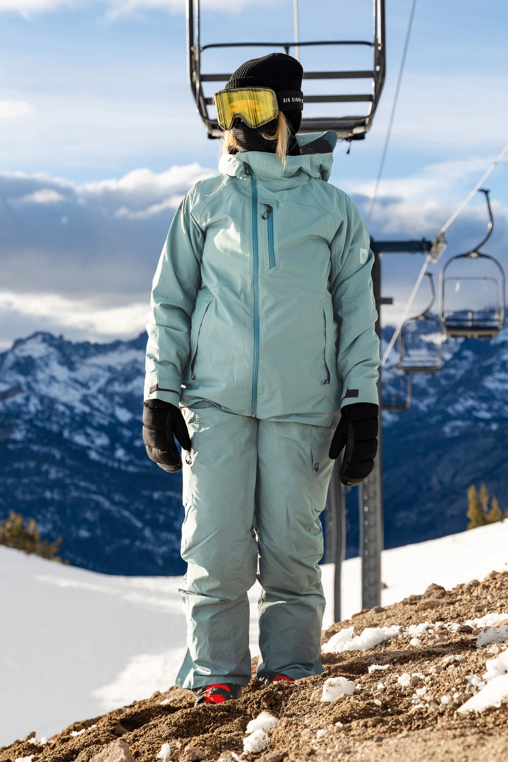 Clad in the 686 Women’s Geode Thermagraph® Pant, along with a light blue snowsuit, black gloves, a black beanie, and yellow ski goggles, a person stands on a snowy slope near a ski lift with mountains in the background—demonstrating the comfort and durability of these 686 waterproof pants that are ideal for winter adventures.