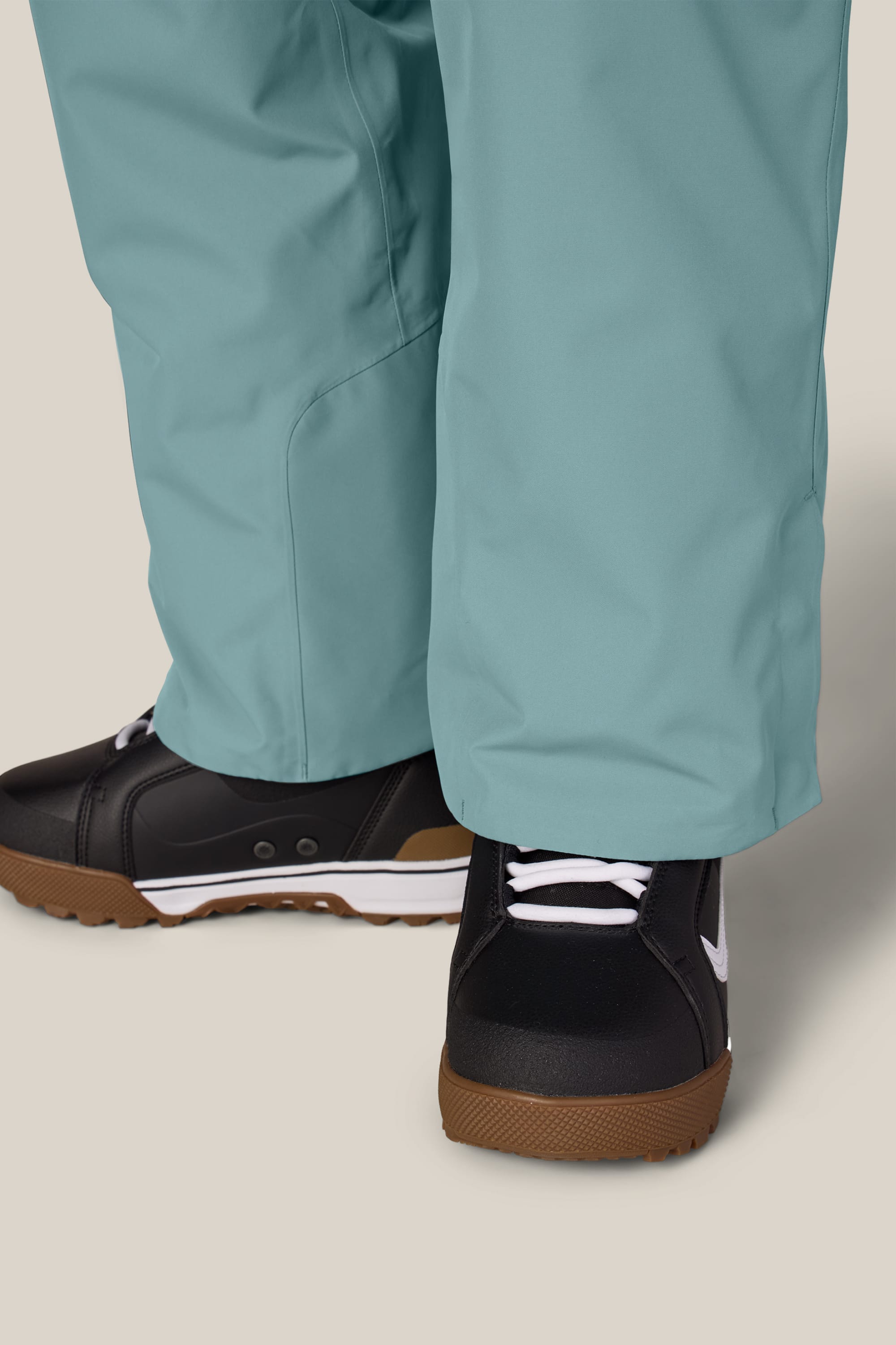 A close-up of a person wearing the 686 Women’s Geode Thermagraph® Pant in teal and black sneakers with white laces and gum soles, standing on a light-colored surface.