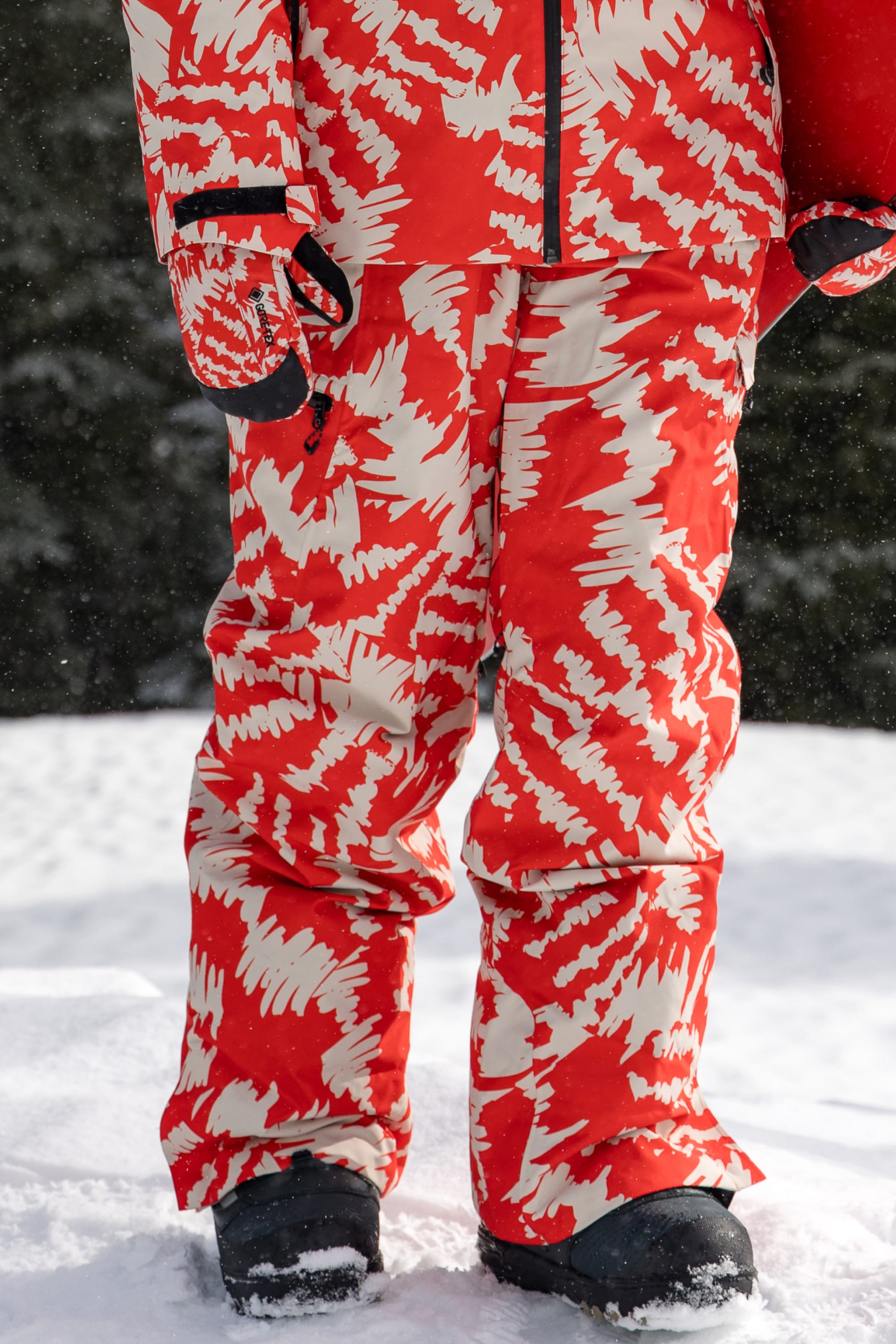 A person stands on snow wearing the 686 Women’s Geode Thermagraph® Pant, paired with a red and beige patterned waterproof jacket and black snow boots. Only the lower half of their body is visible as snowflakes fall against a backdrop of dark trees.