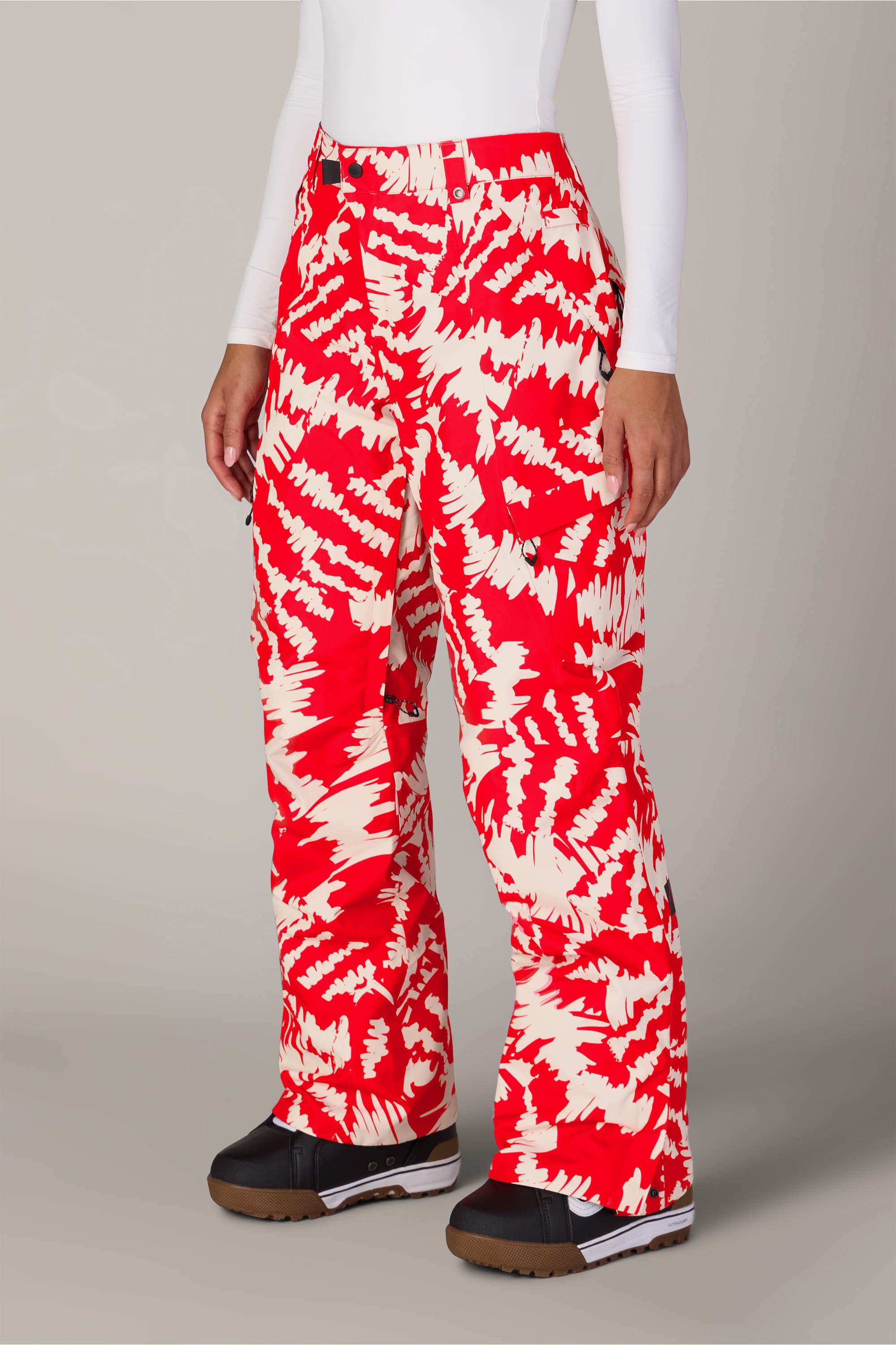 A person wearing the 686 Women’s Geode Thermagraph® Pant by 686—red and white patterned women’s waterproof pants—paired with a white long-sleeve top and black boots, standing against a plain light background.