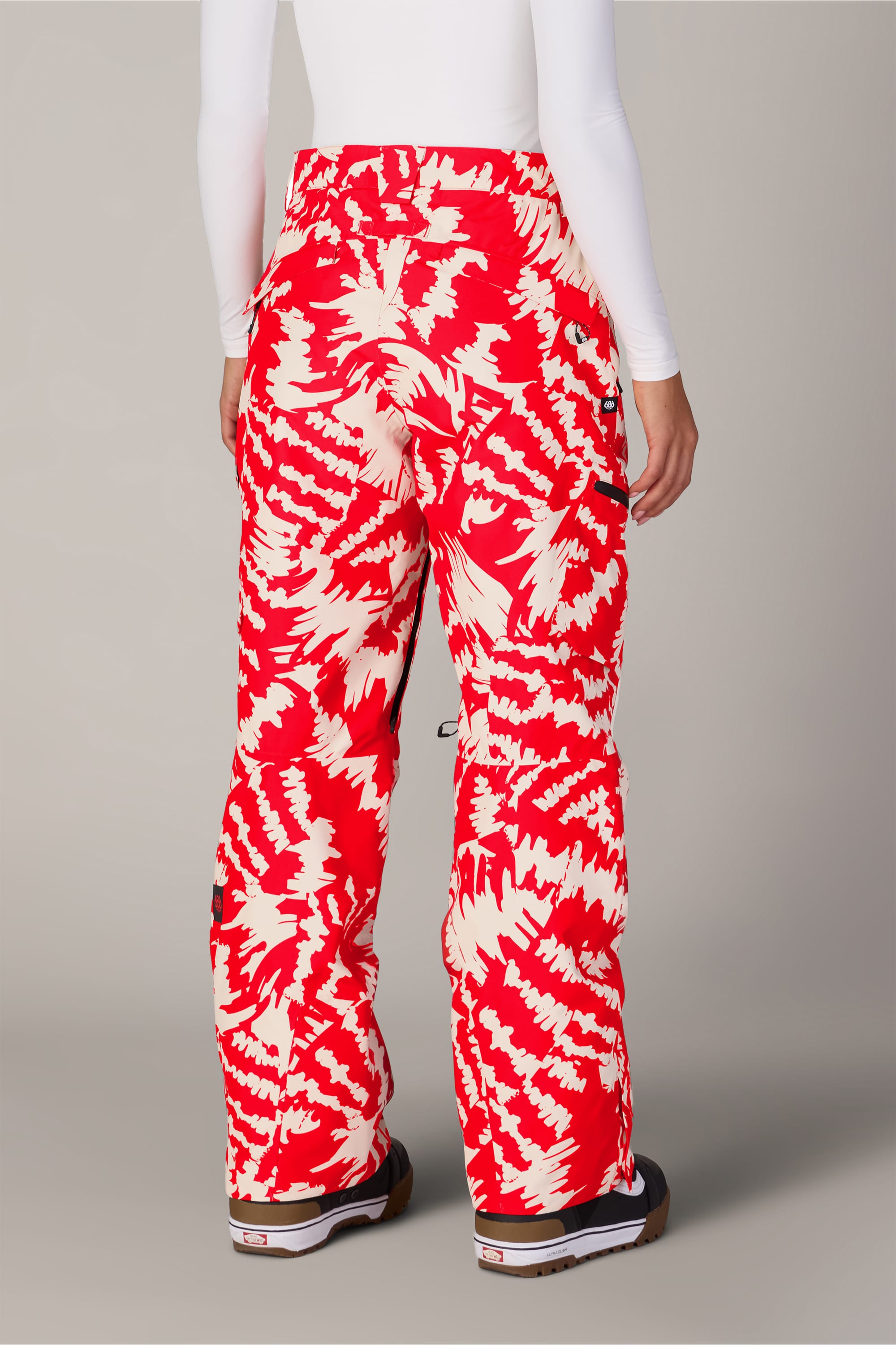 A person wearing 686 Women’s Geode Thermagraph® Pant by 686, which features a red and white pattern, paired with a white long-sleeve shirt and black-and-white shoes, stands facing away from the camera against a plain background.