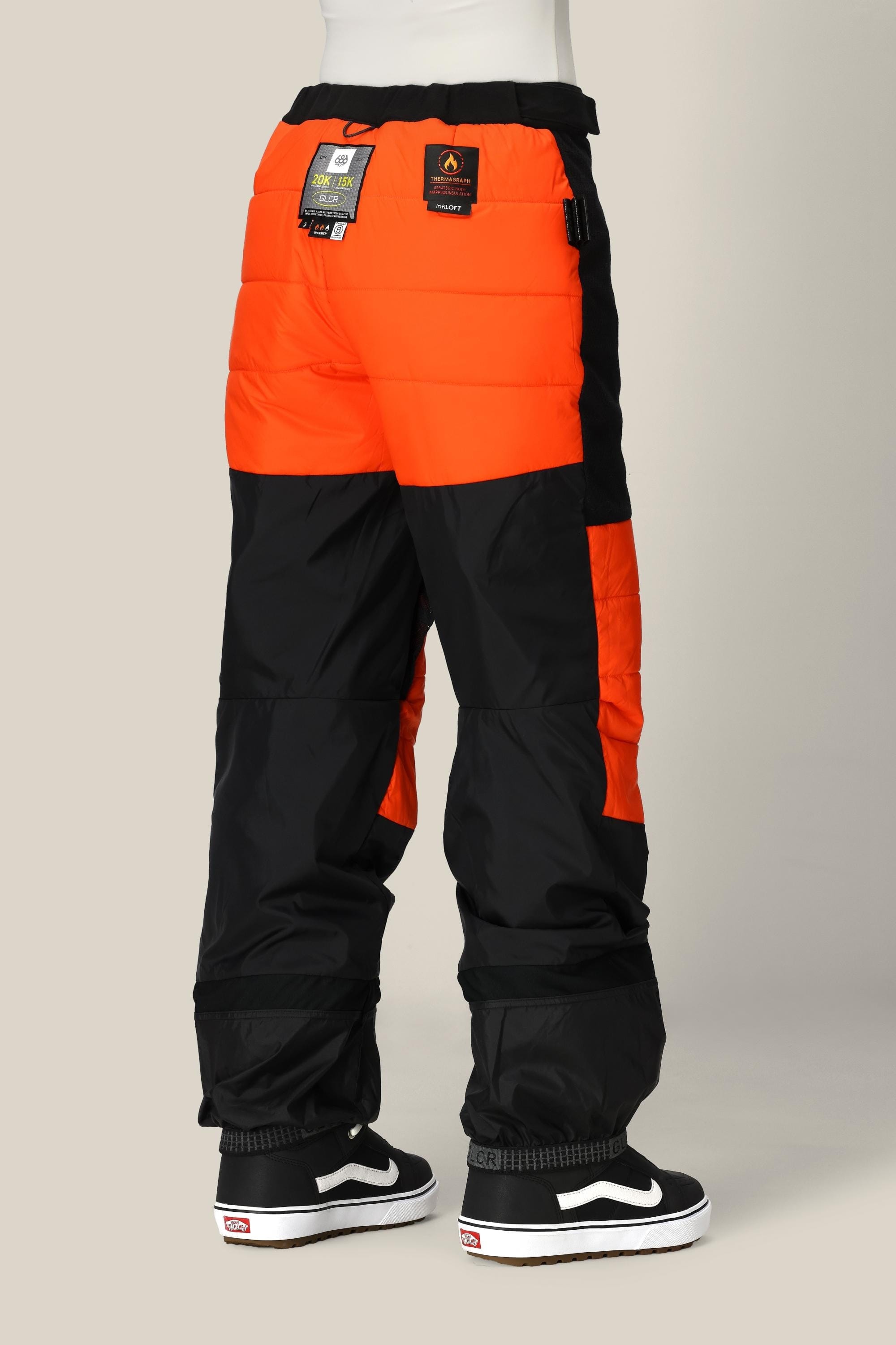 A person is wearing the 686 Women’s Geode Thermagraph® Pant in bright orange and black, with tags attached, standing against a neutral background. These pants by 686 are crafted from waterproof polyester for style and functionality. The outfit is completed with black and white Vans sneakers.