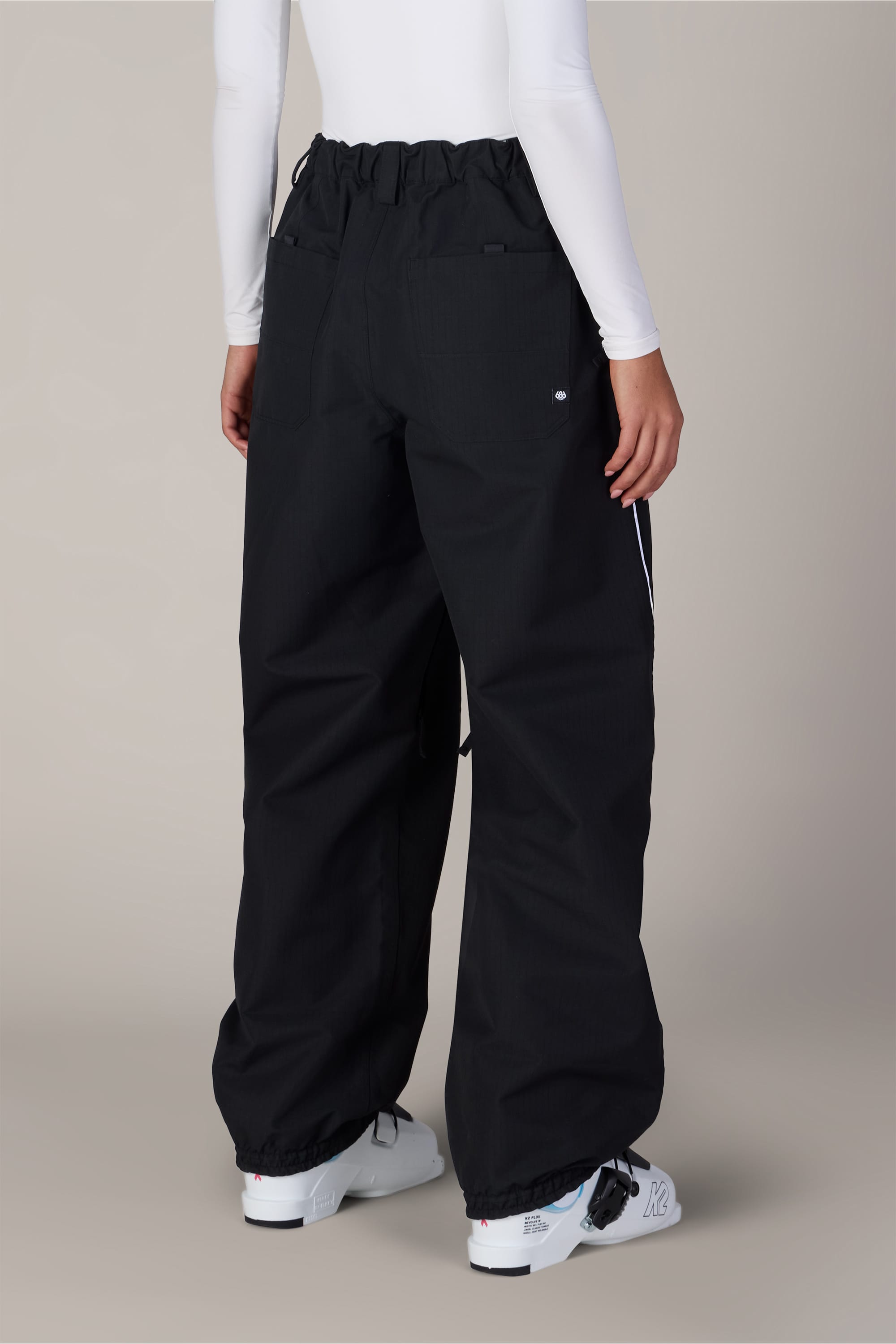 A person shown from the back against a plain background is wearing the 686 Women’s Hellkat™ Shell Pant in black, paired with a white long-sleeve top and white sneakers.