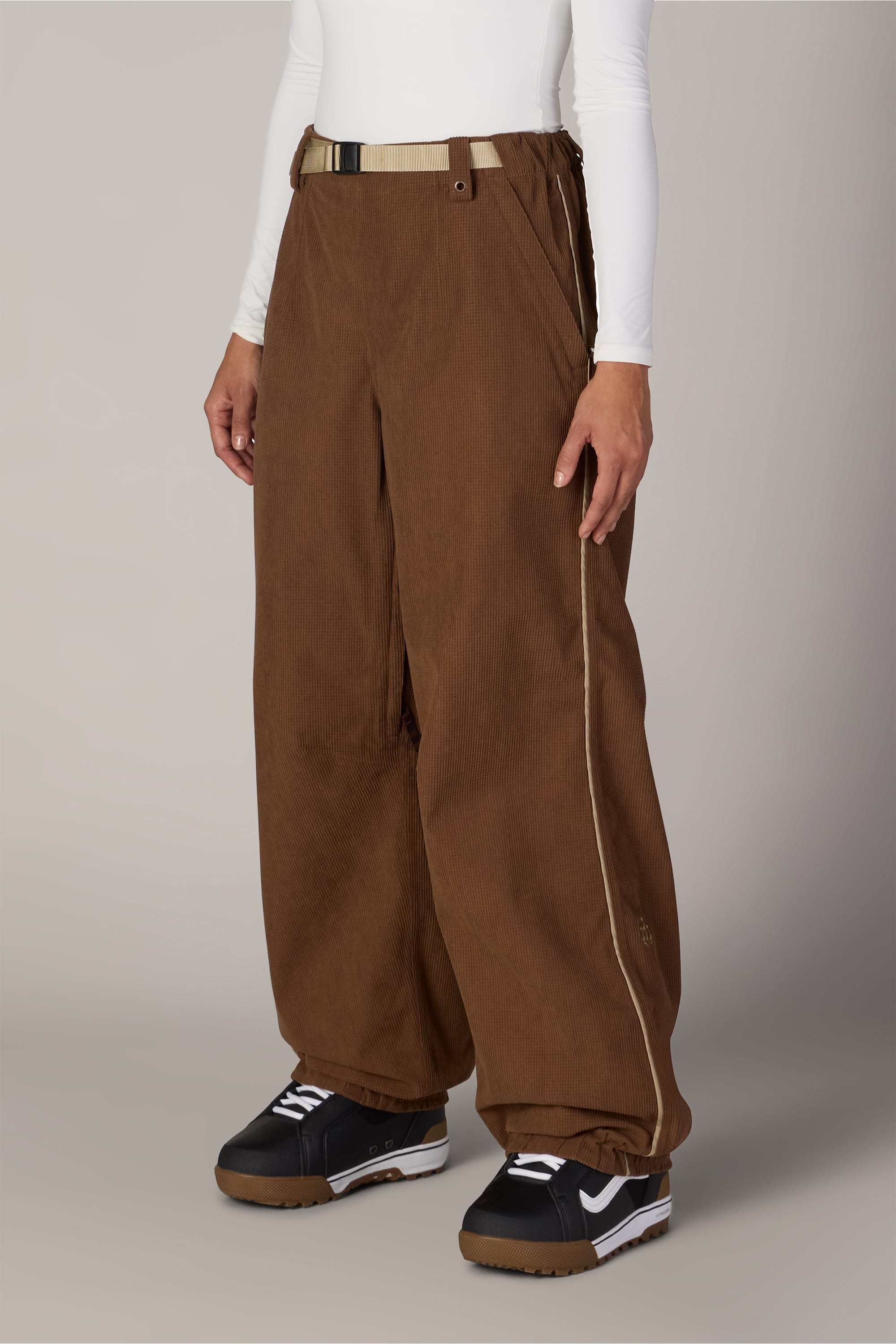A person is wearing the 686 Women’s Hellkat™ Shell Pant by 686—loose brown corduroy women’s pants with a wide-leg fit and cream side piping—styled with a beige belt, white long-sleeve top, and black-and-white athletic shoes, standing against a neutral background.