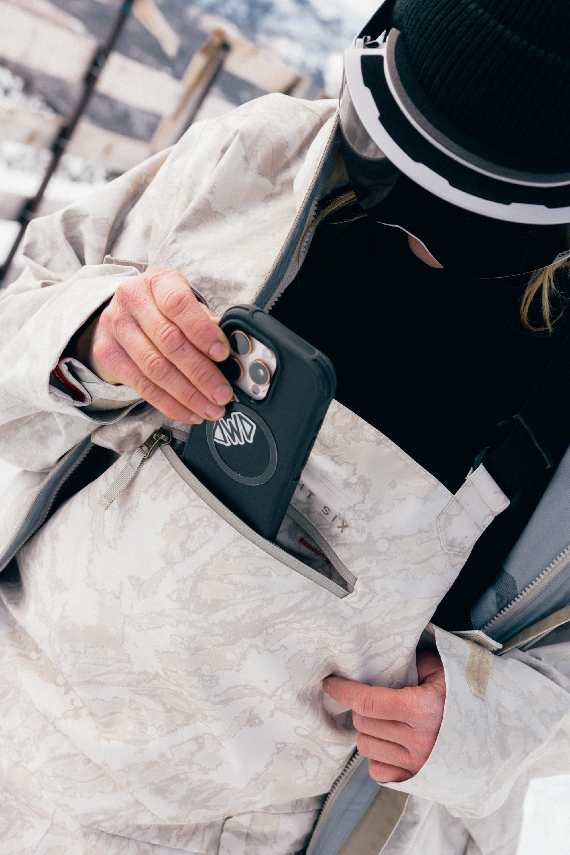 Wearing the 686 Women’s Regalia™ Bib, a person in winter gear and goggles slides their phone into the front pocket of the waterproof jacket, with snowy mountains visible in the background.