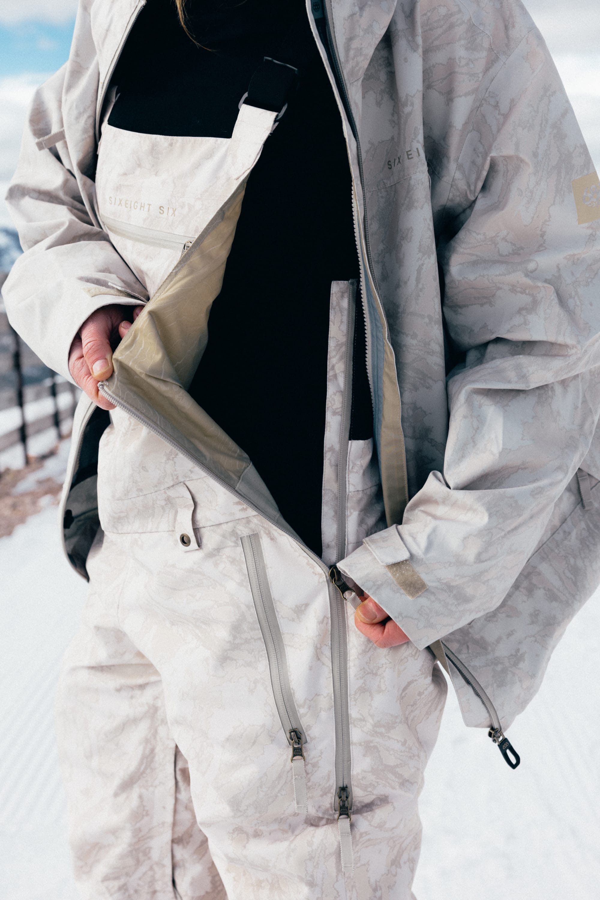 A person wearing light-colored winter gear unzips their snow jacket, revealing a black 686 Women’s Regalia™ Bib underneath. Snow and a fence are visible in the background.