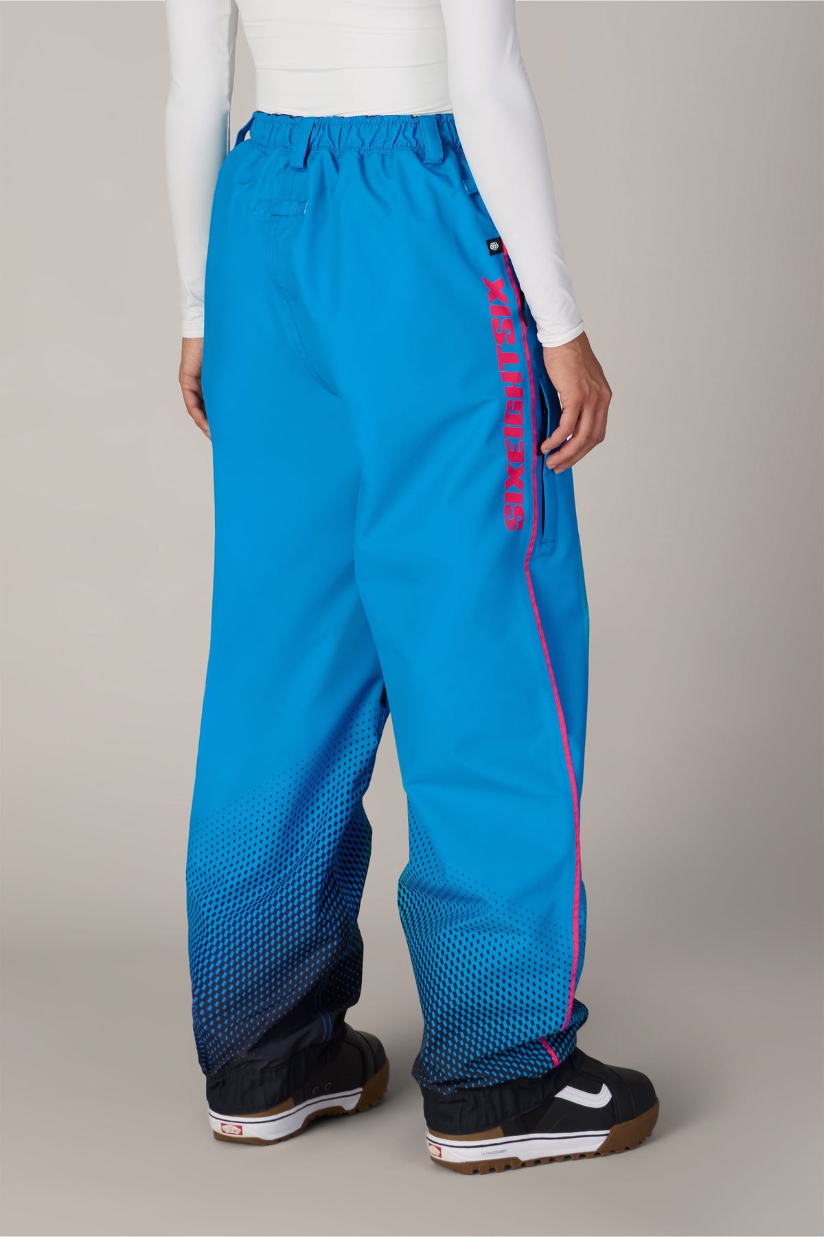 A person wearing bright blue 686 Women’s Fox Racing 97 Shell Pants with a pink 686 logo on the side and a black dotted gradient pattern at the cuffs.